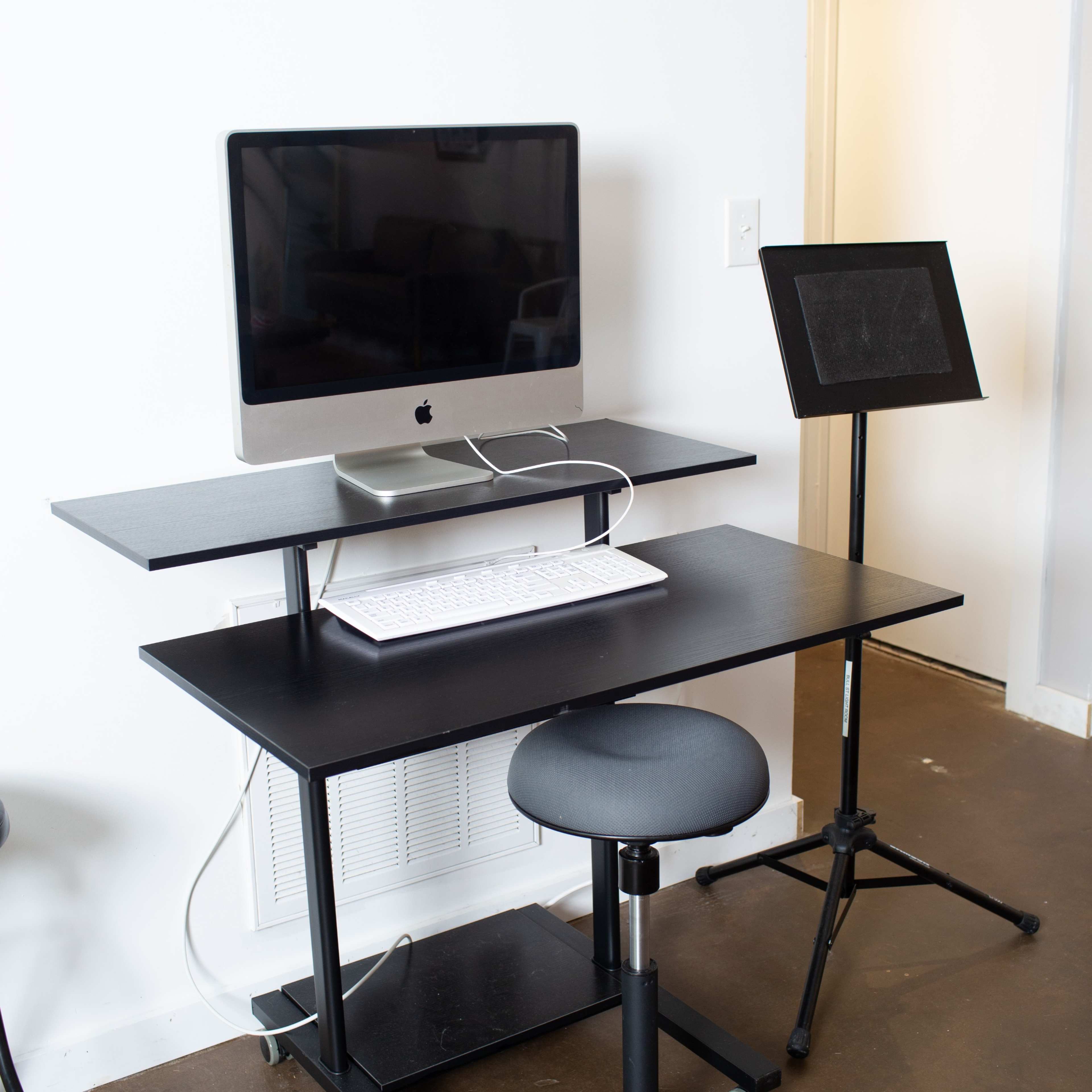 A modern workspace featuring a black desk with a computer monitor, laptop, and keyboard, accompanied by a stool and an adjustable music stand.