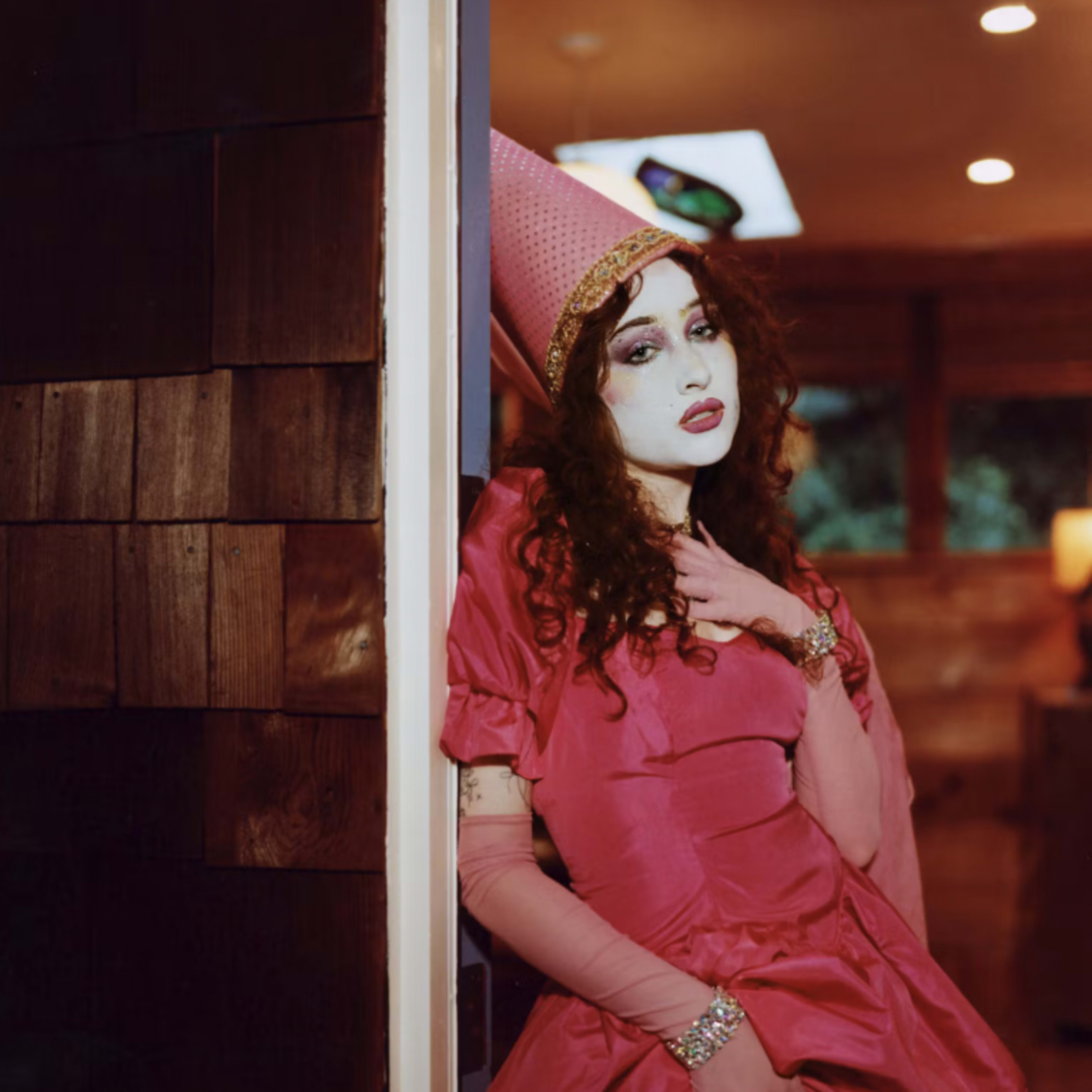 A person in a pink dress and a large pink hat stands by a doorway, posing dramatically in a modern interior space.