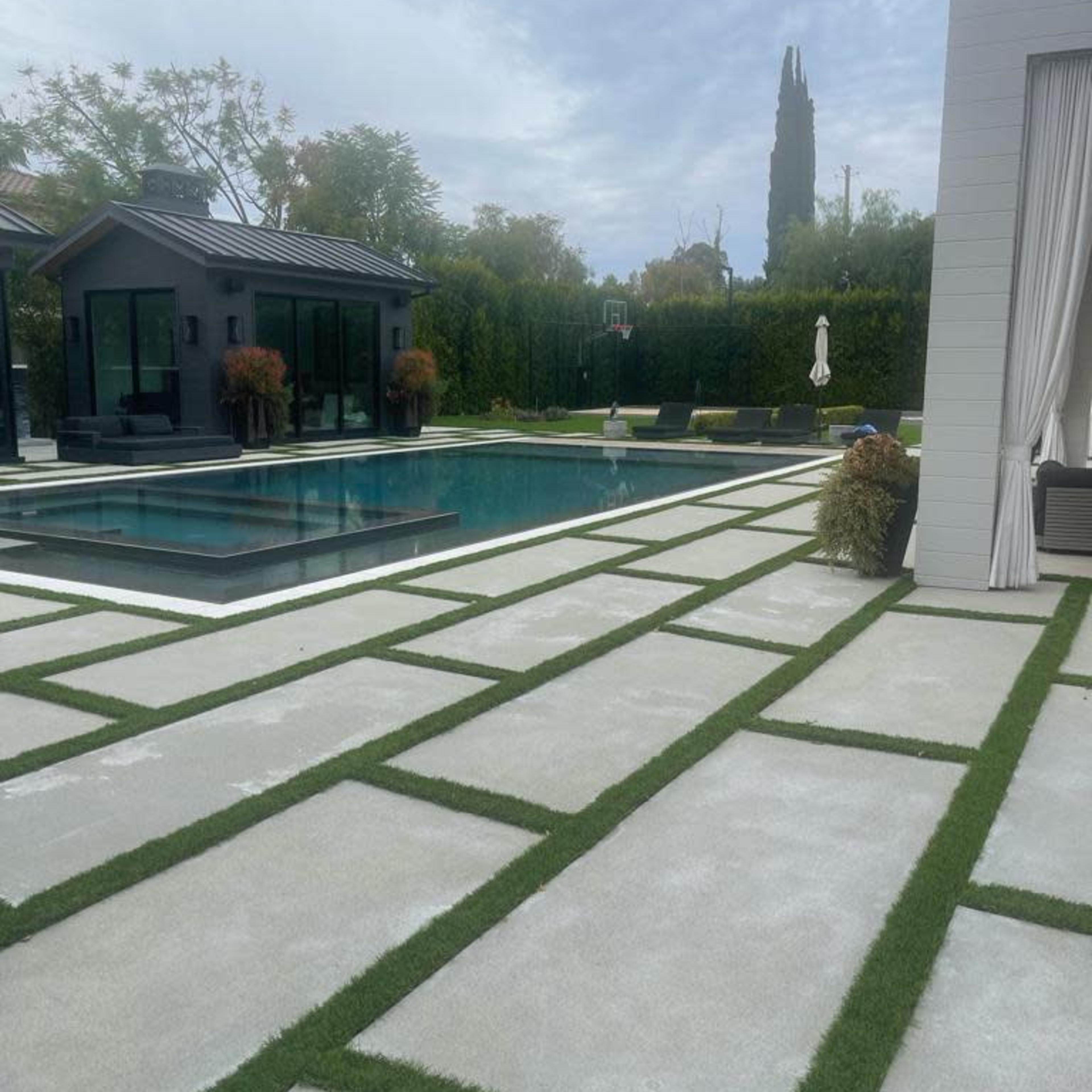 A modern outdoor space featuring a swimming pool, a small building with large windows, and a patio area with rectangular concrete slabs and patches of grass.