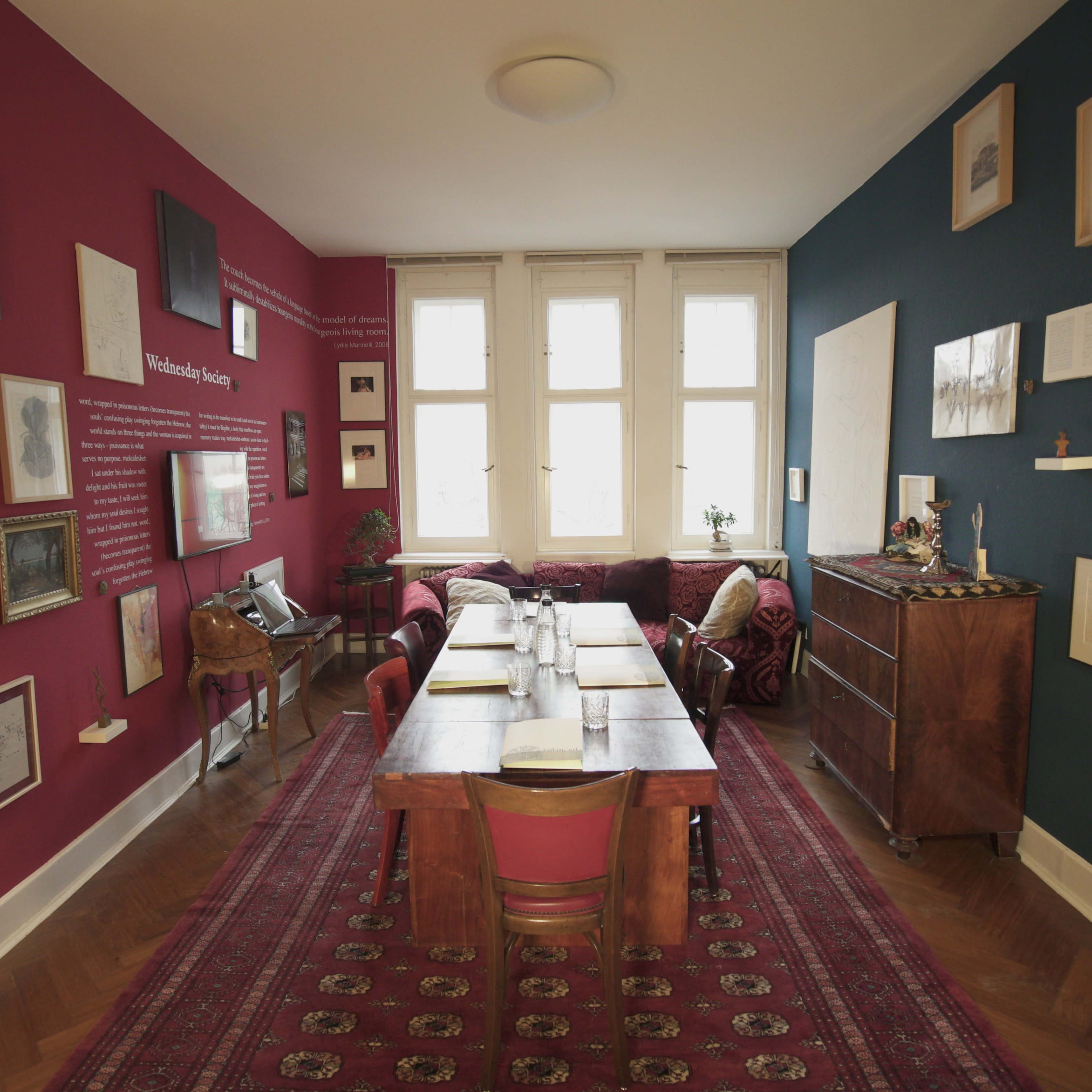 A well-decorated dining room with a long wooden table surrounded by chairs, against a backdrop of red and blue walls adorned with framed artwork and photographs.
