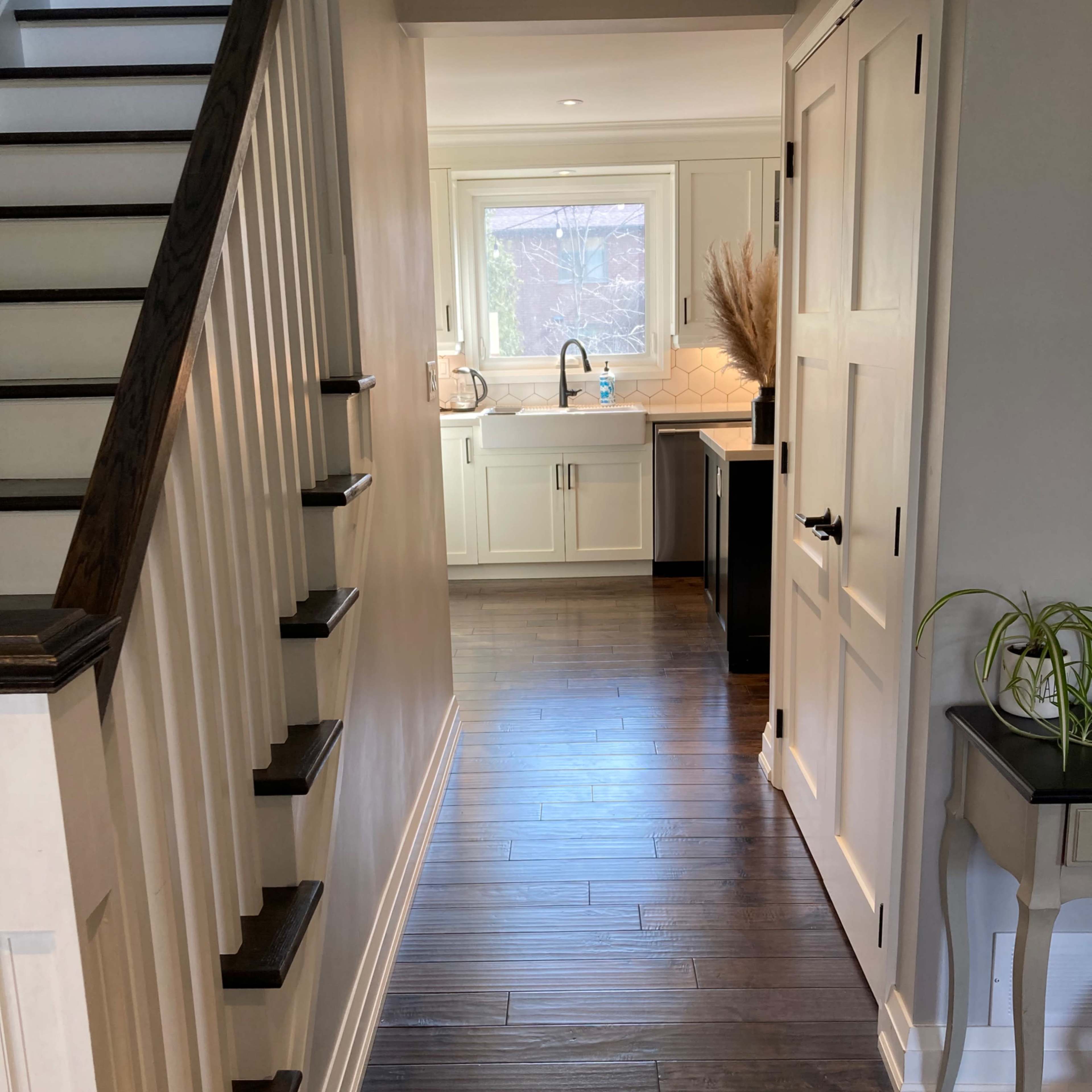 A hallway with wooden flooring leads to a well-lit kitchen, visible at the end, with a staircase on the left and a closed door on the right.