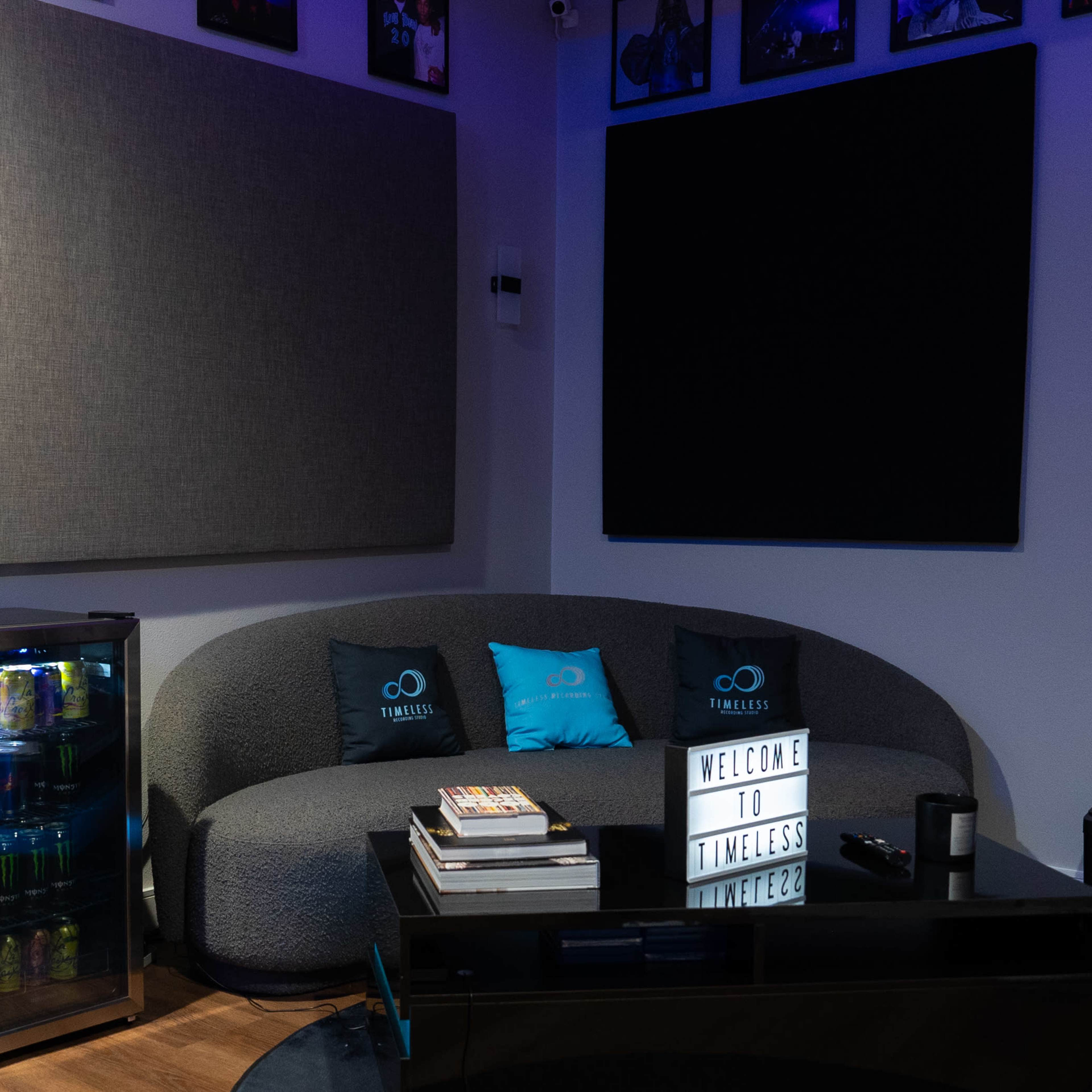 A modern lounge area with a curved gray couch, a glass coffee table stacked with books, and a small beverage fridge beside it, all set against a backdrop of framed pictures on the walls.