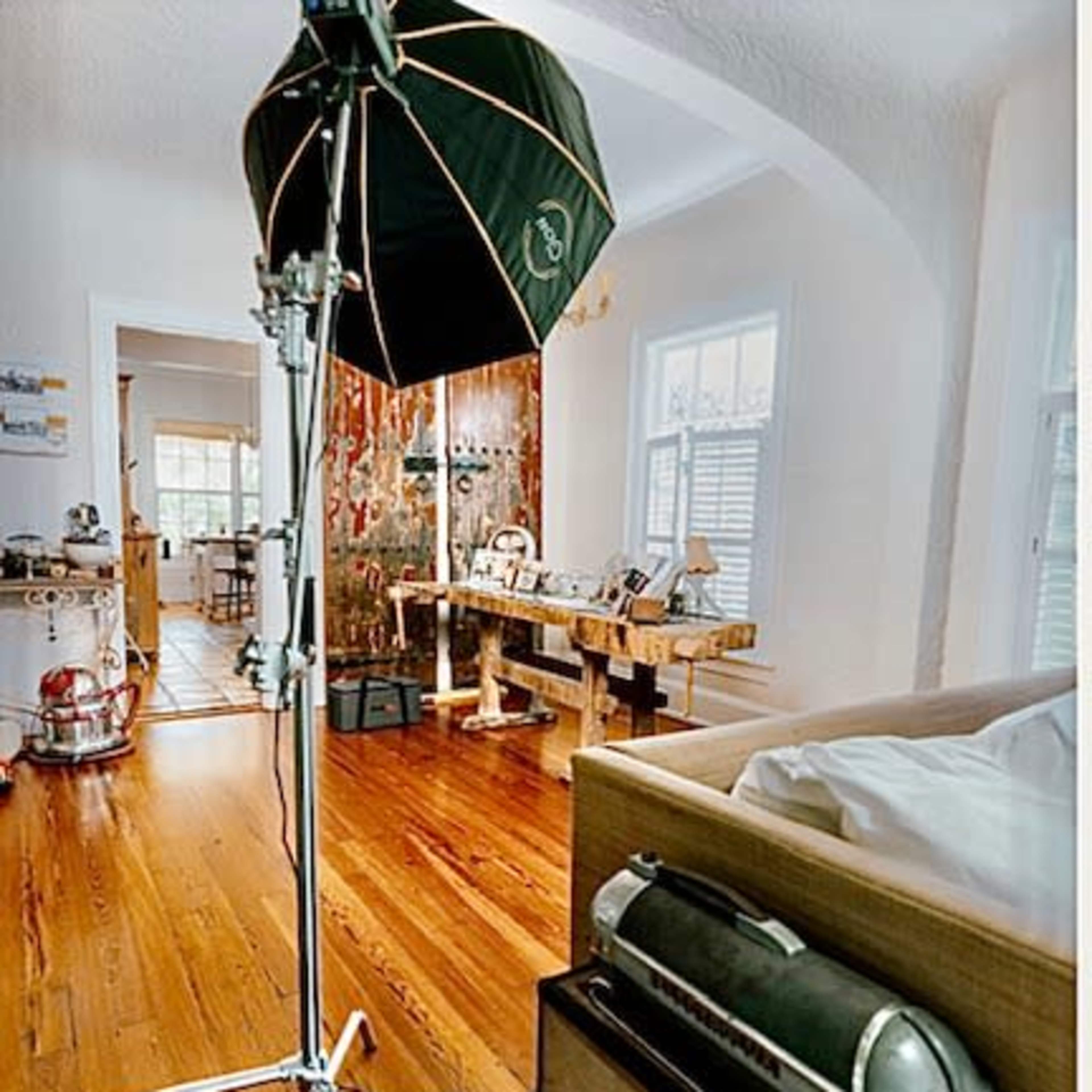A photography lighting setup stands beside a bed, casting light on a living space with wooden floors and a wooden table in the background.