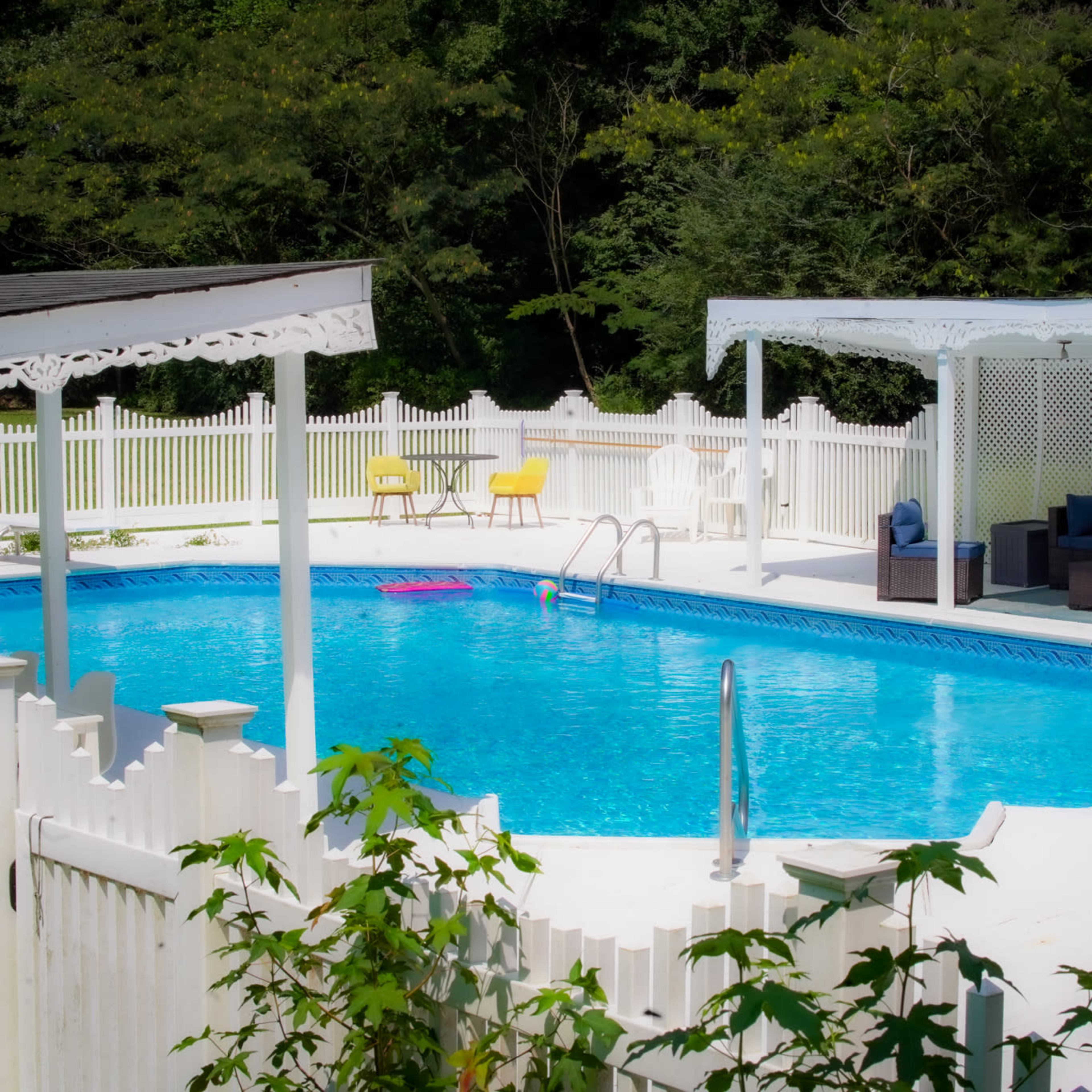 A rectangular swimming pool surrounded by a white fence and lounge area featuring outdoor furniture and shaded seating.