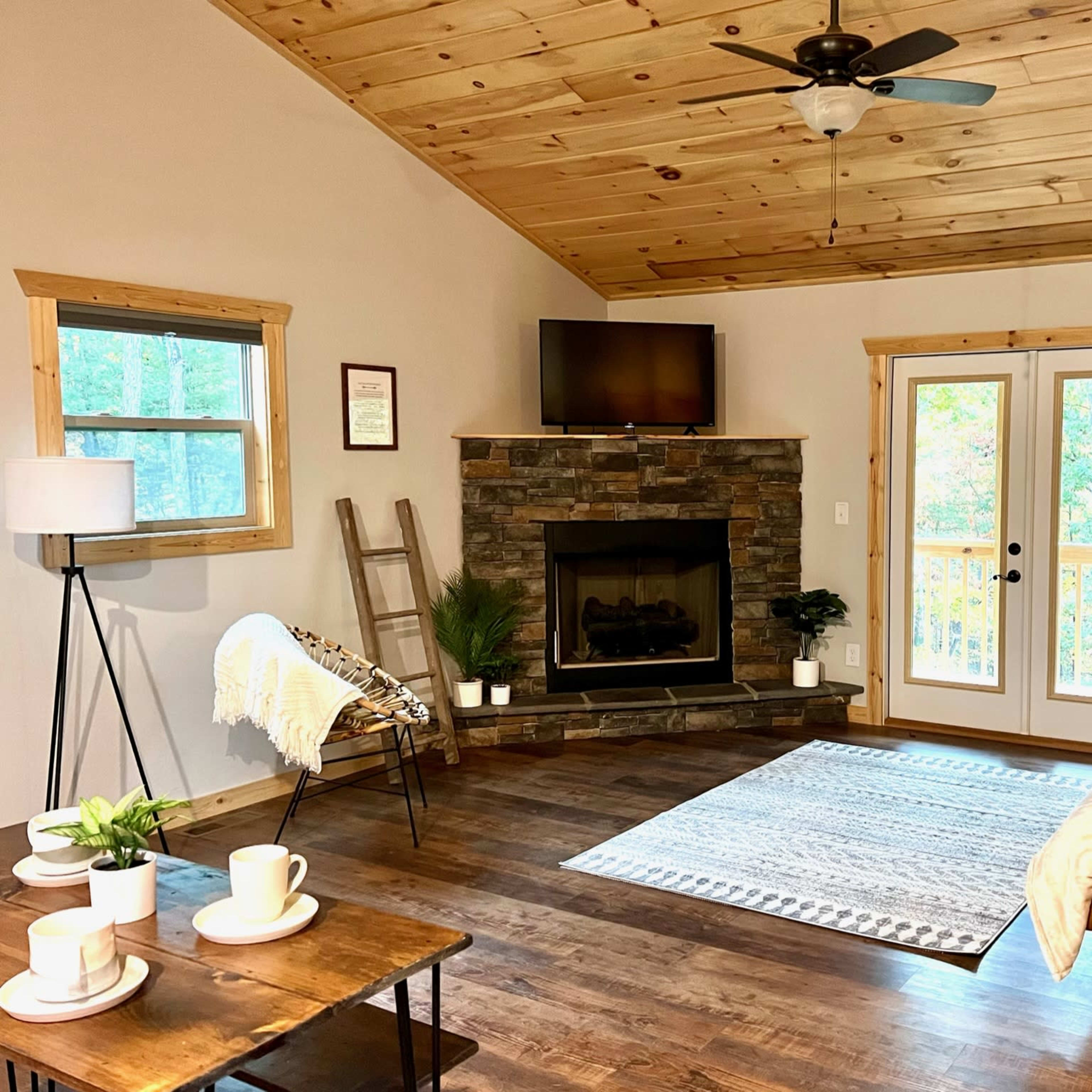 A cozy living room with a stone fireplace, a wooden ceiling, a brown leather sofa, and a large window with a door leading outside.