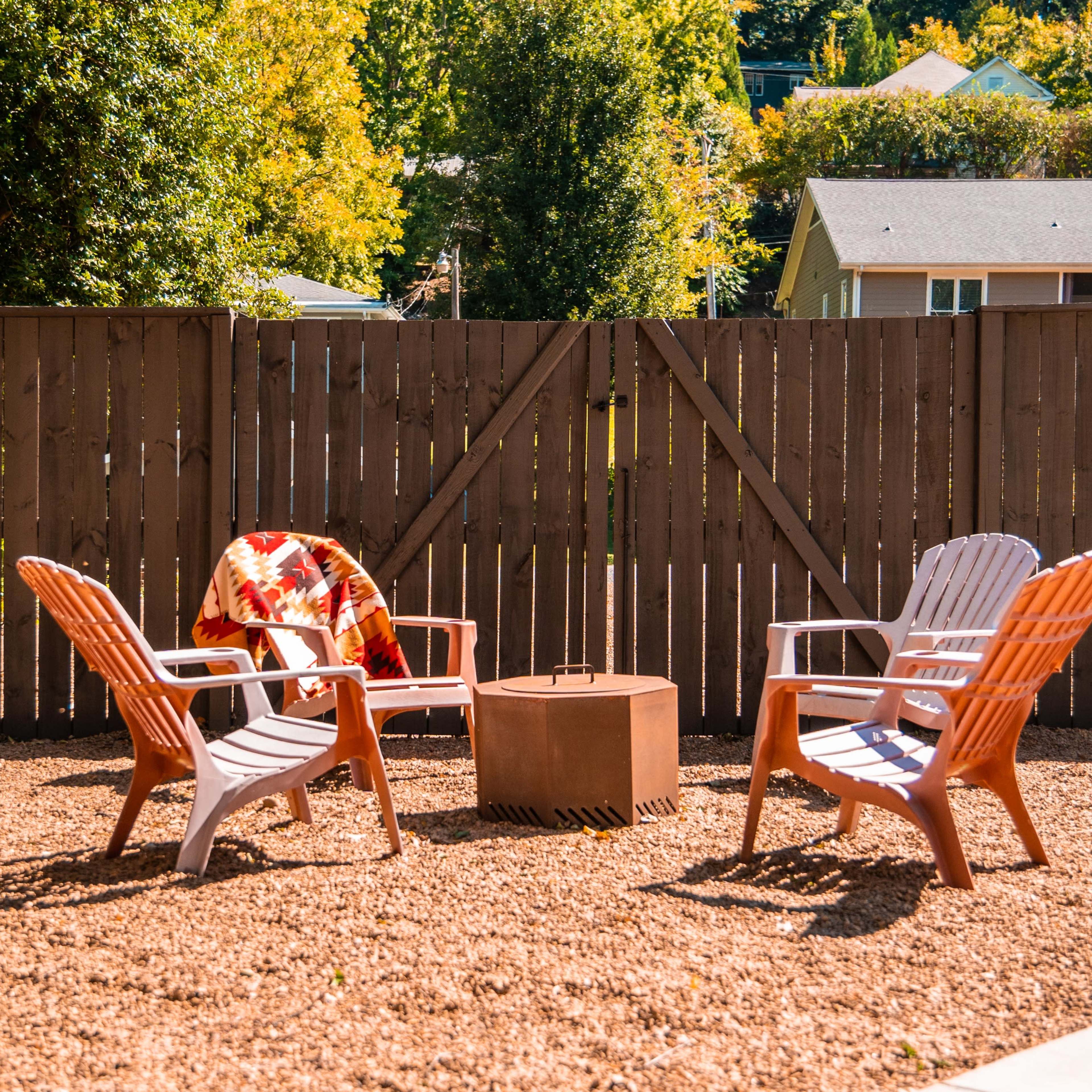A fenced outdoor space with two Adirondack chairs, a small fire pit, and a blanket draped over one chair amidst gravel landscaping.