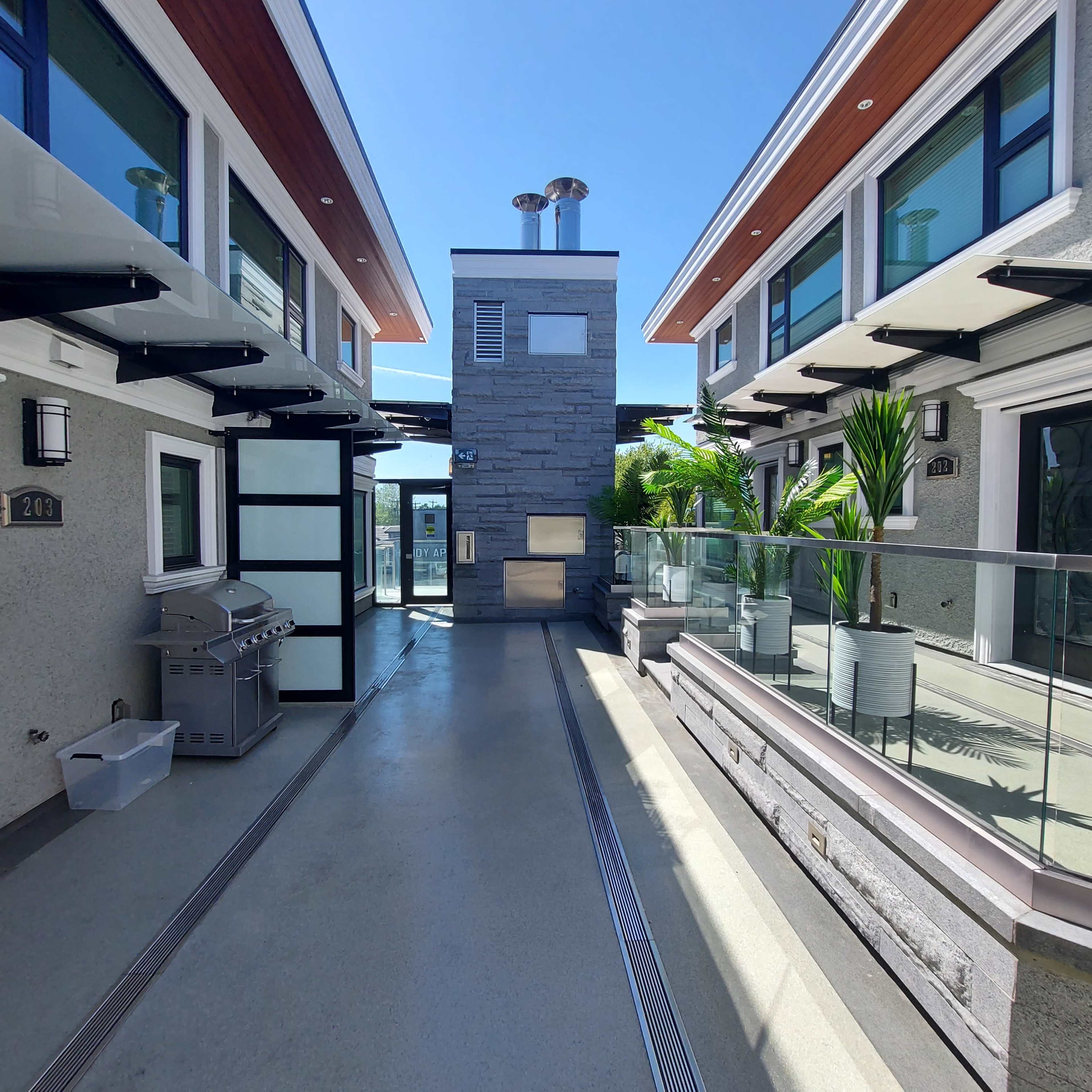 A modern outdoor corridor between two buildings featuring a central stone fireplace, a grill, and potted plants.
