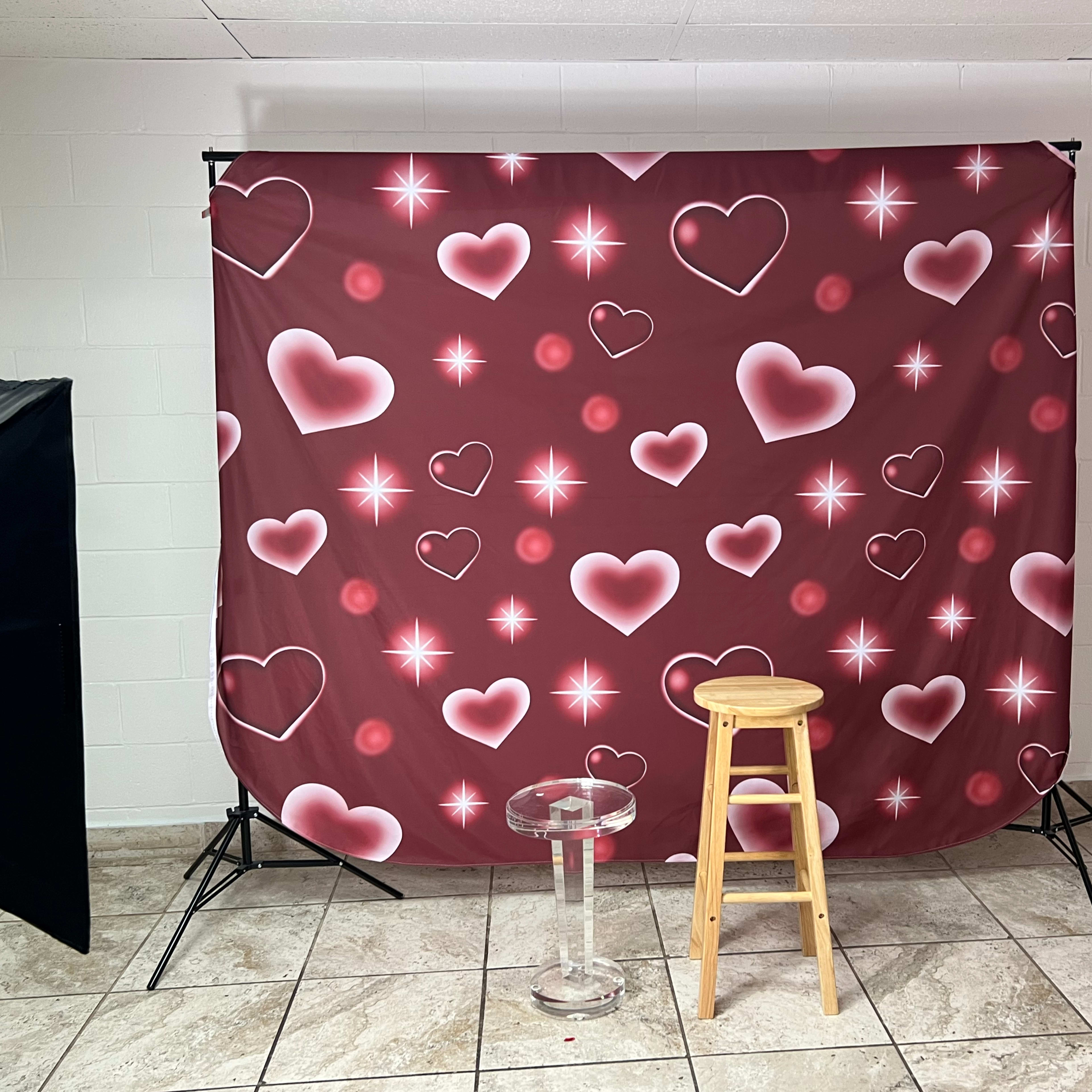 A photography backdrop featuring a pattern of hearts and stars in shades of pink and red, set up in a room with a stool and a glass table in front of it.