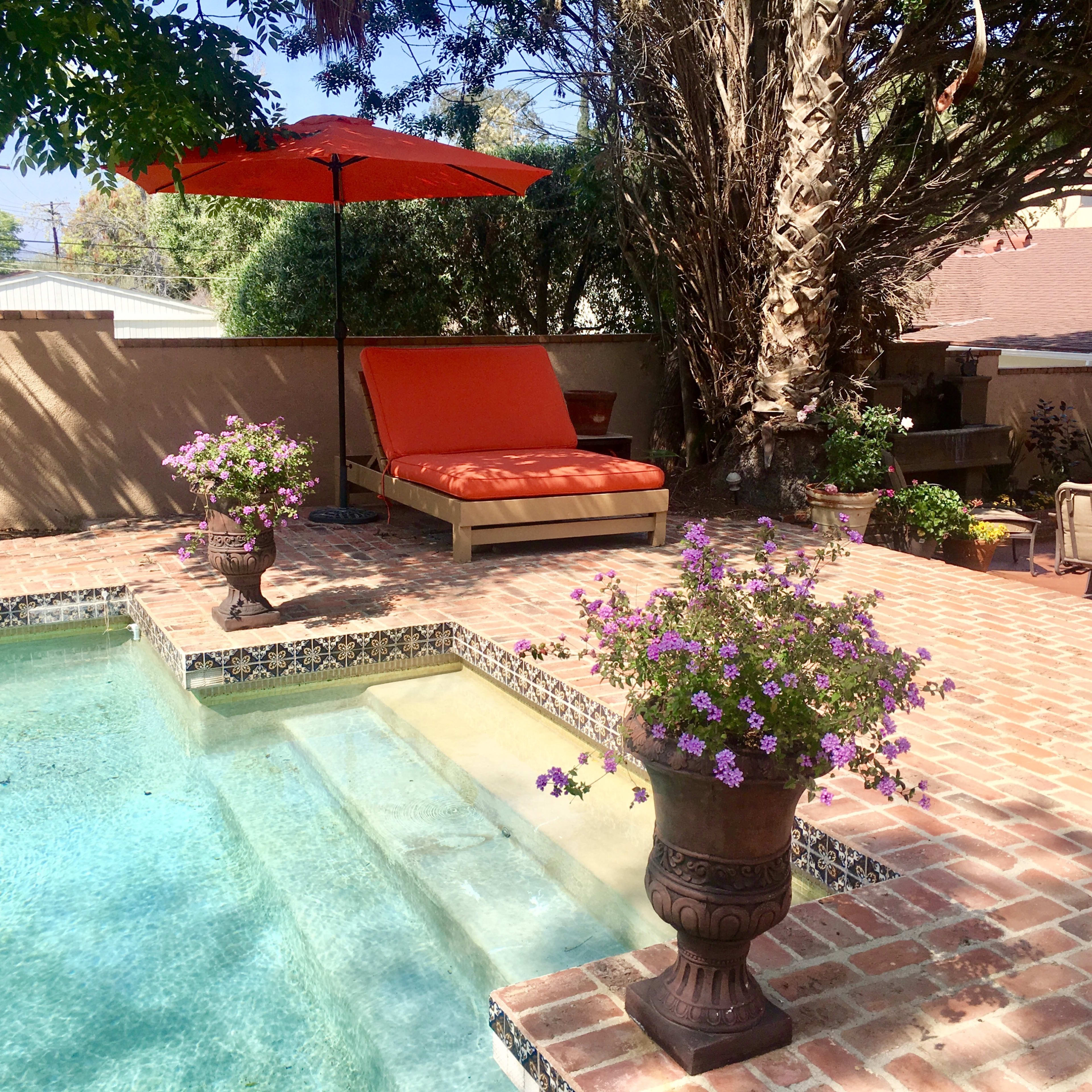 A backyard pool area featuring a lounge chair under an orange umbrella, surrounded by potted flowers and brick patio.