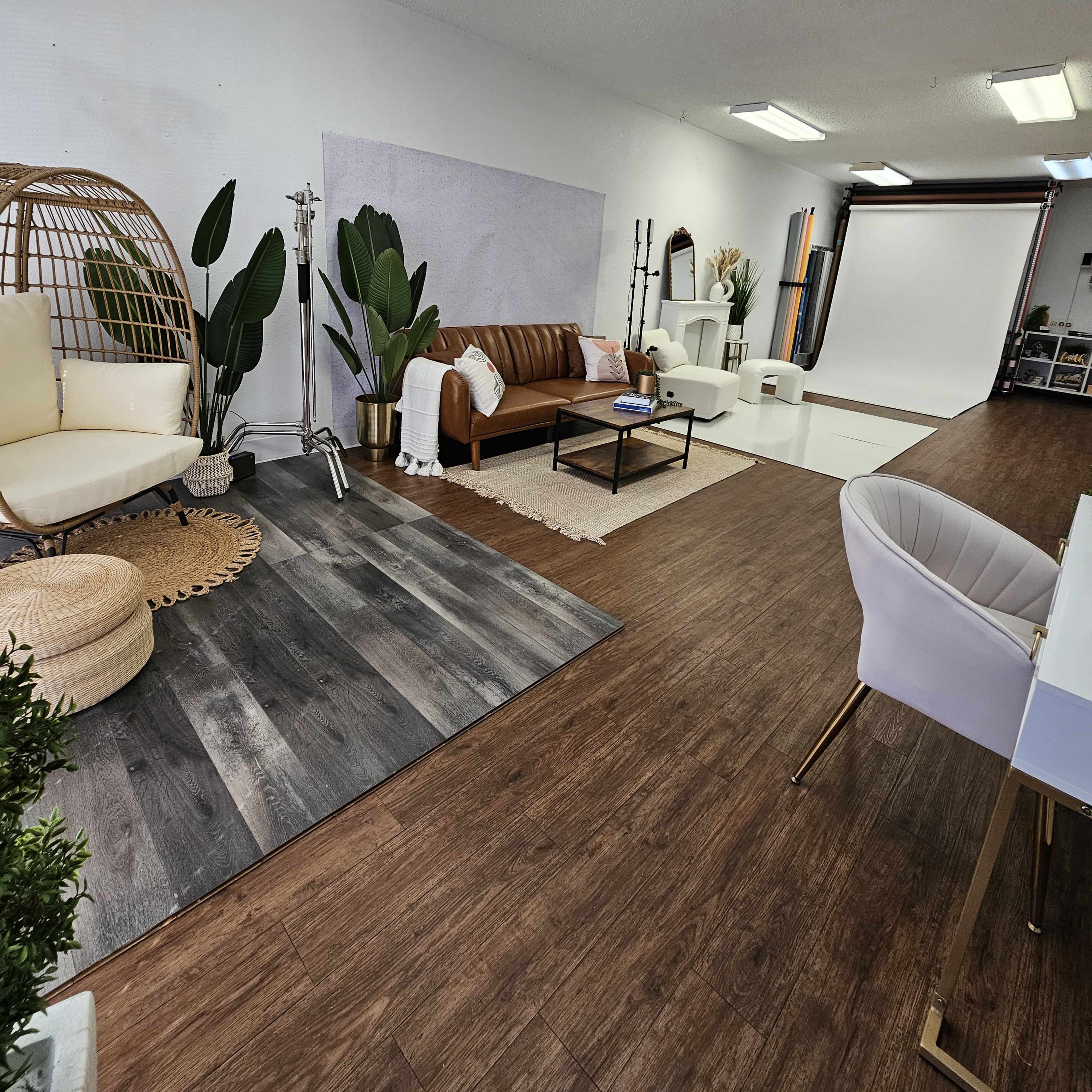 A spacious photography studio featuring a brown leather sofa, a light-colored hanging chair, large plants, and a backdrop area with studio lighting equipment.