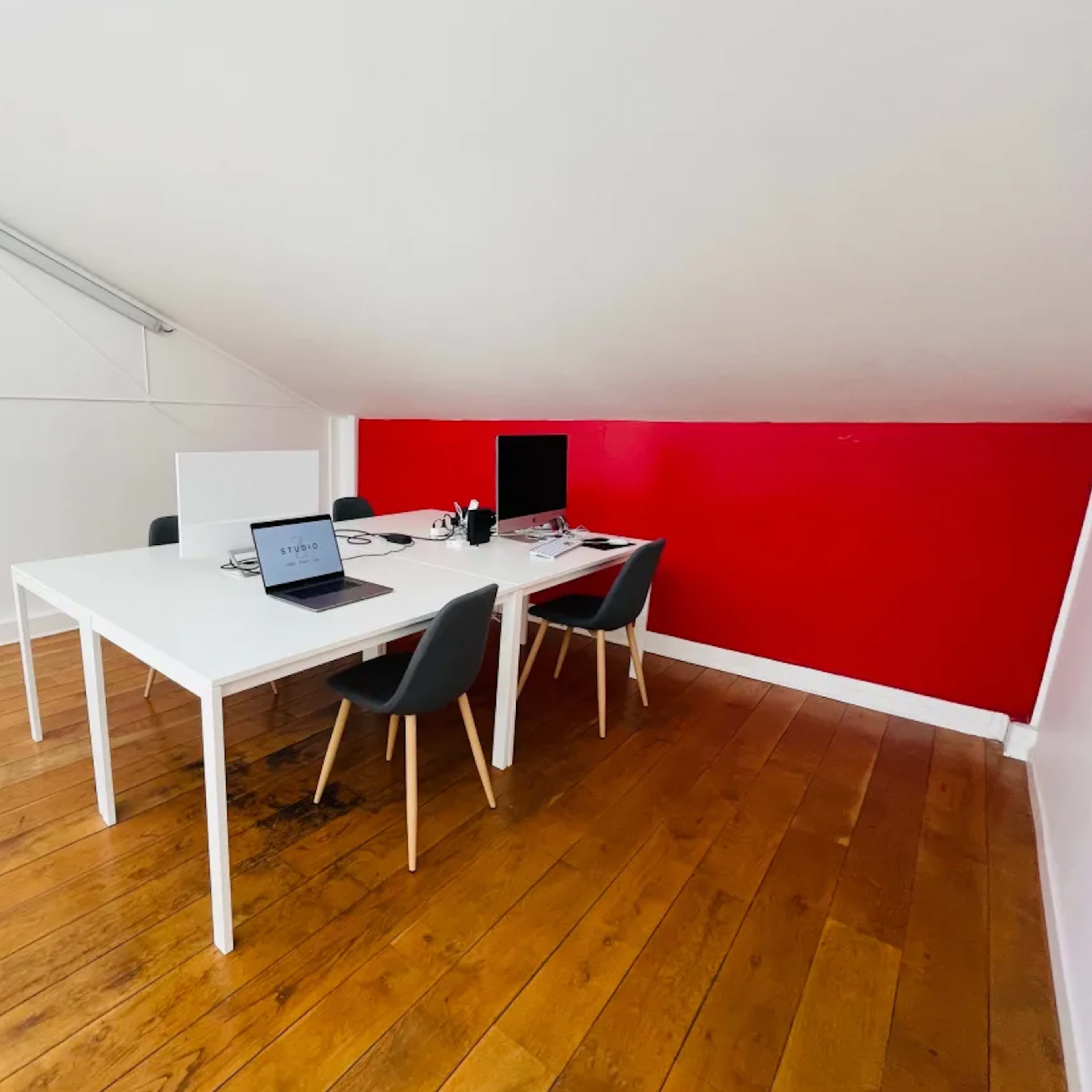 A minimalist office space with two desks, one laptop, and a monitor, set against a red accent wall.