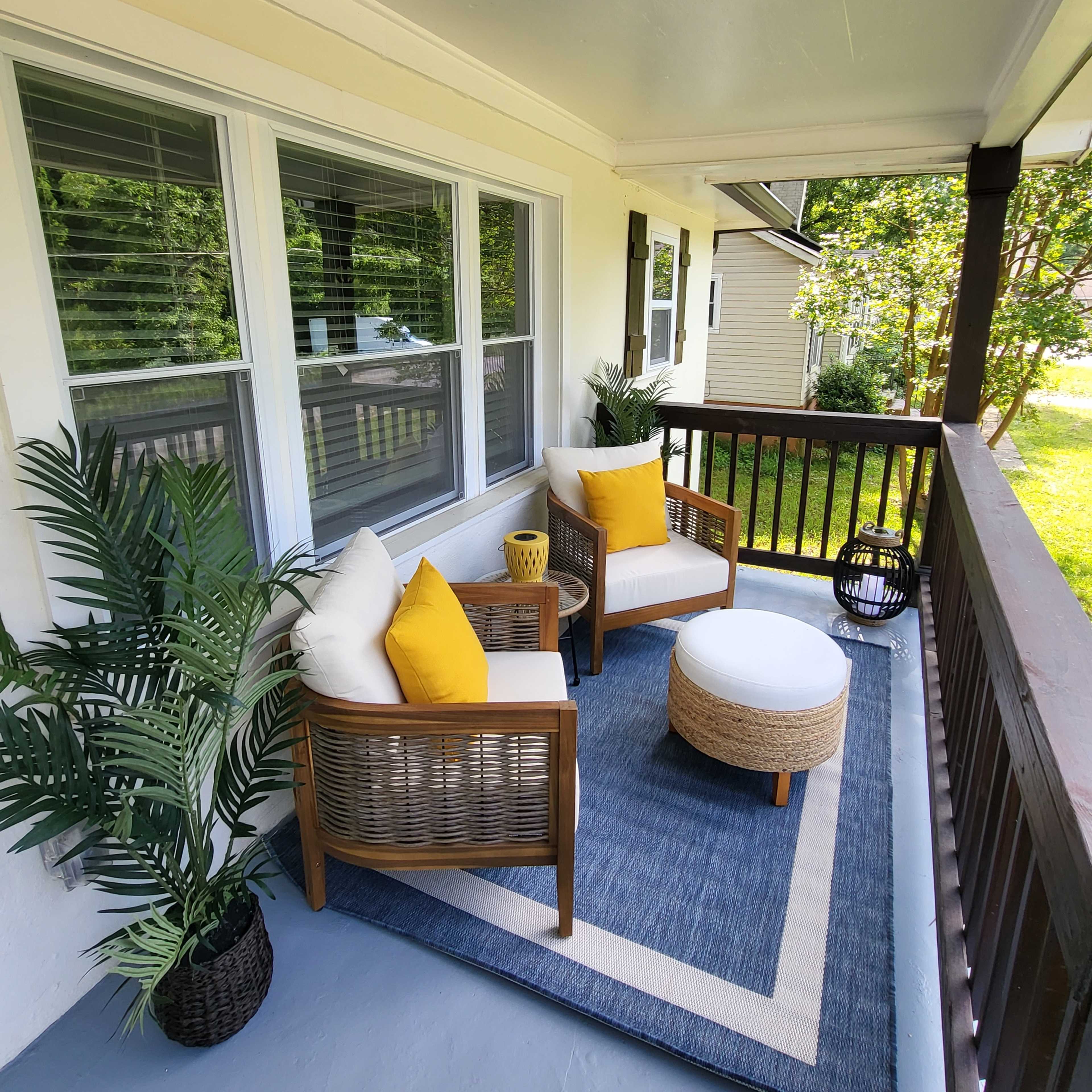 A porch featuring two wicker chairs with yellow pillows, a round ottoman, and a patterned rug, surrounded by potted plants.