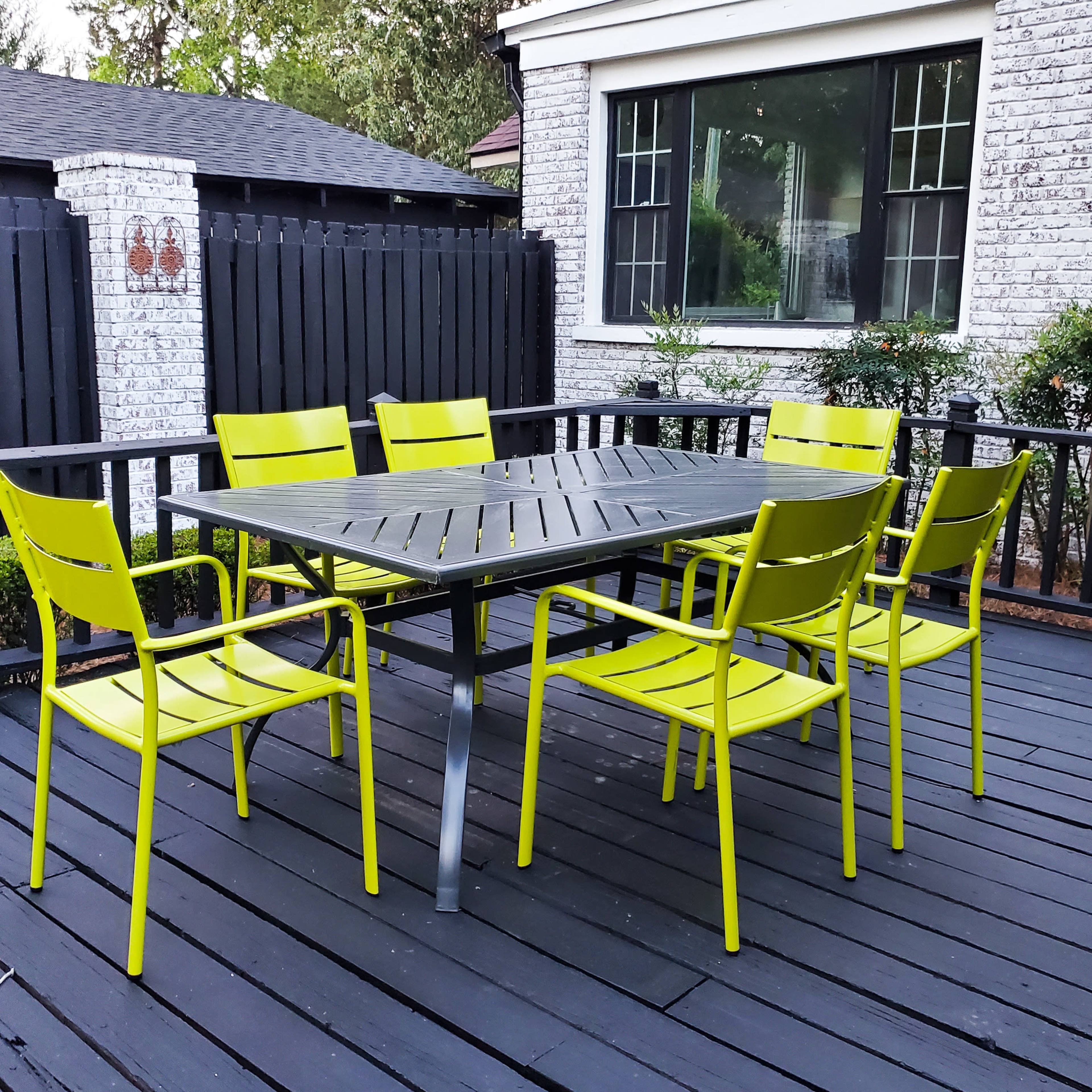A rectangular table with six bright yellow chairs is arranged on a dark wooden deck next to a house.