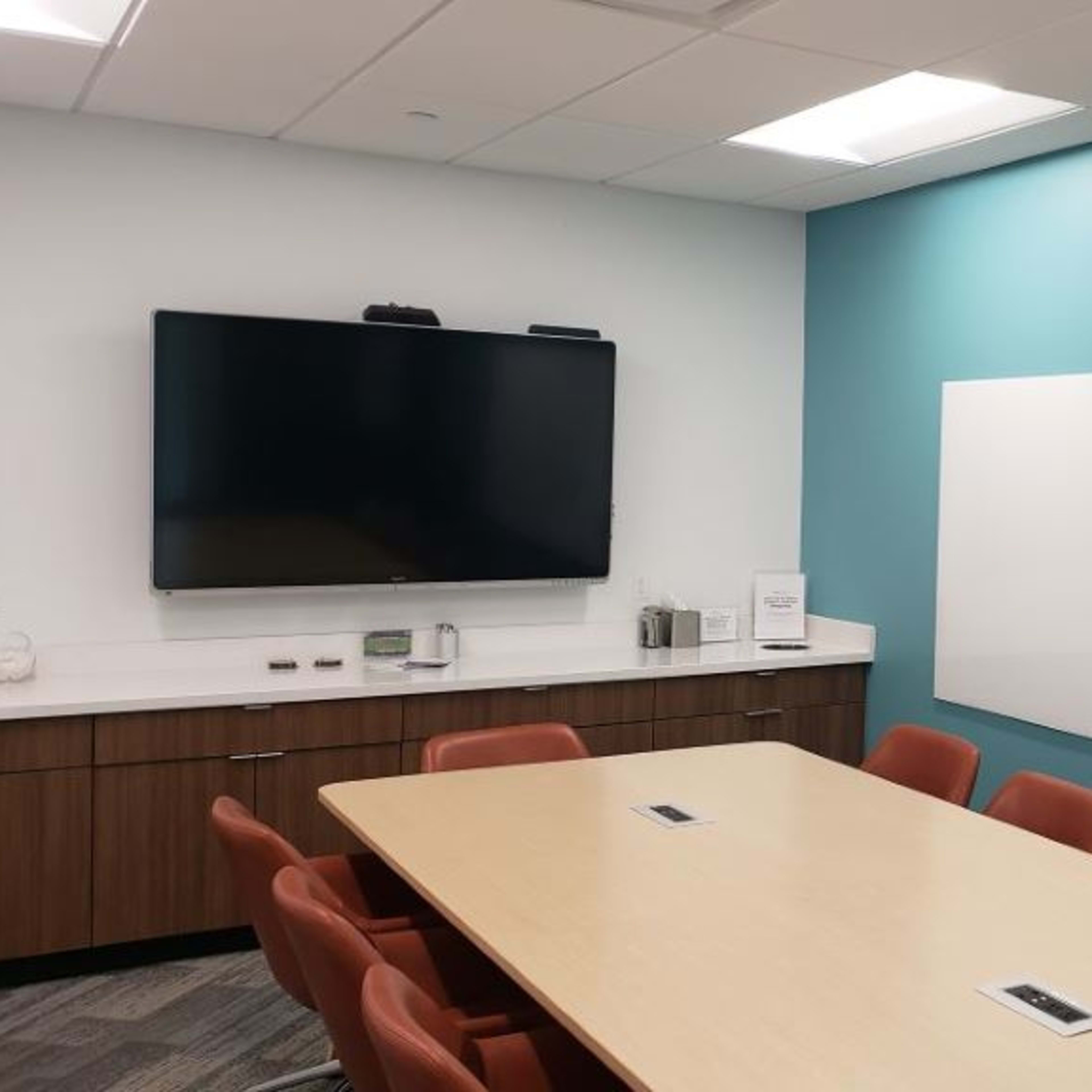 A conference room features a large table surrounded by orange chairs, a television mounted on the wall, and two whiteboards.