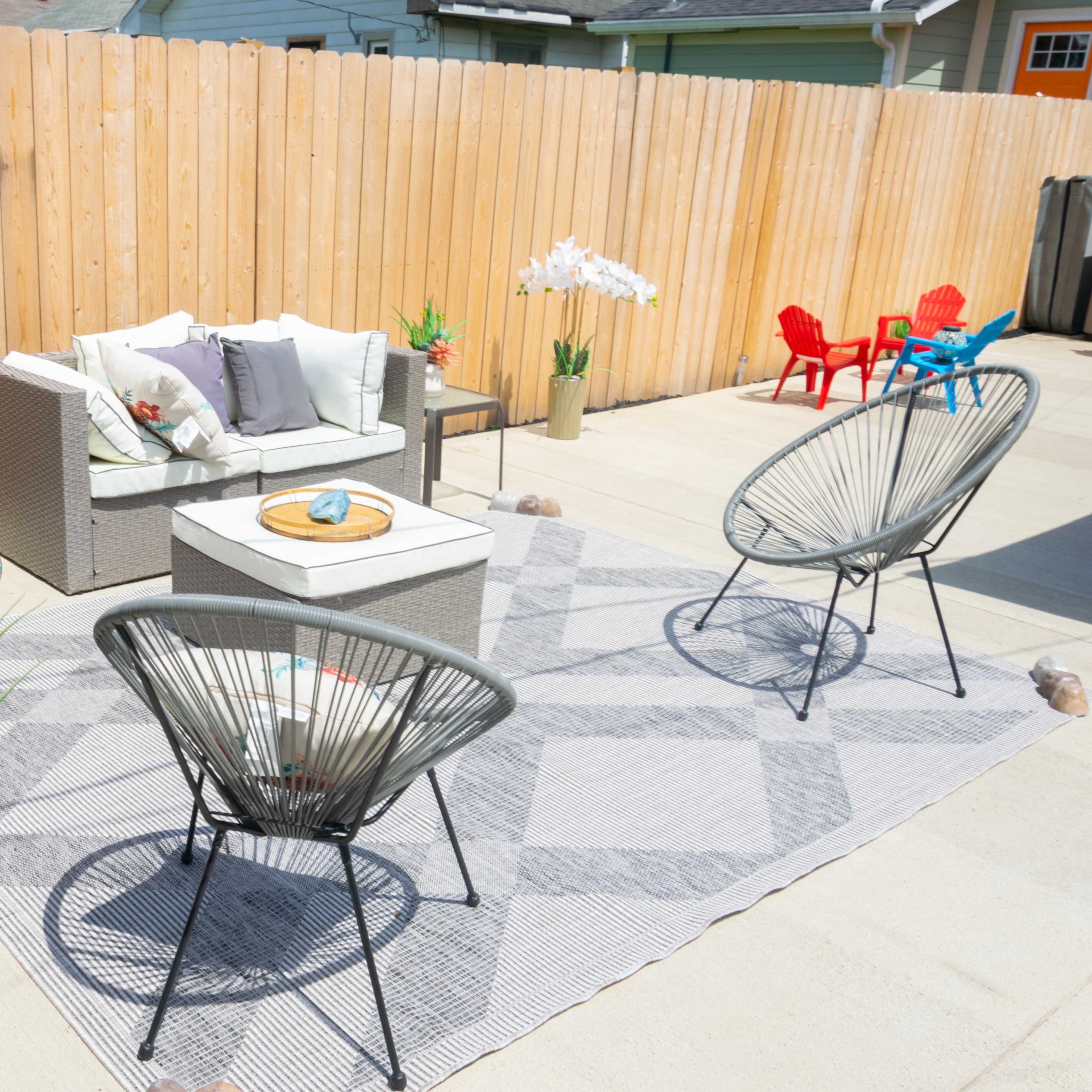 A patio area features a sectional sofa, two circular chairs, decorative plants, and a central rug, surrounded by a wooden fence.