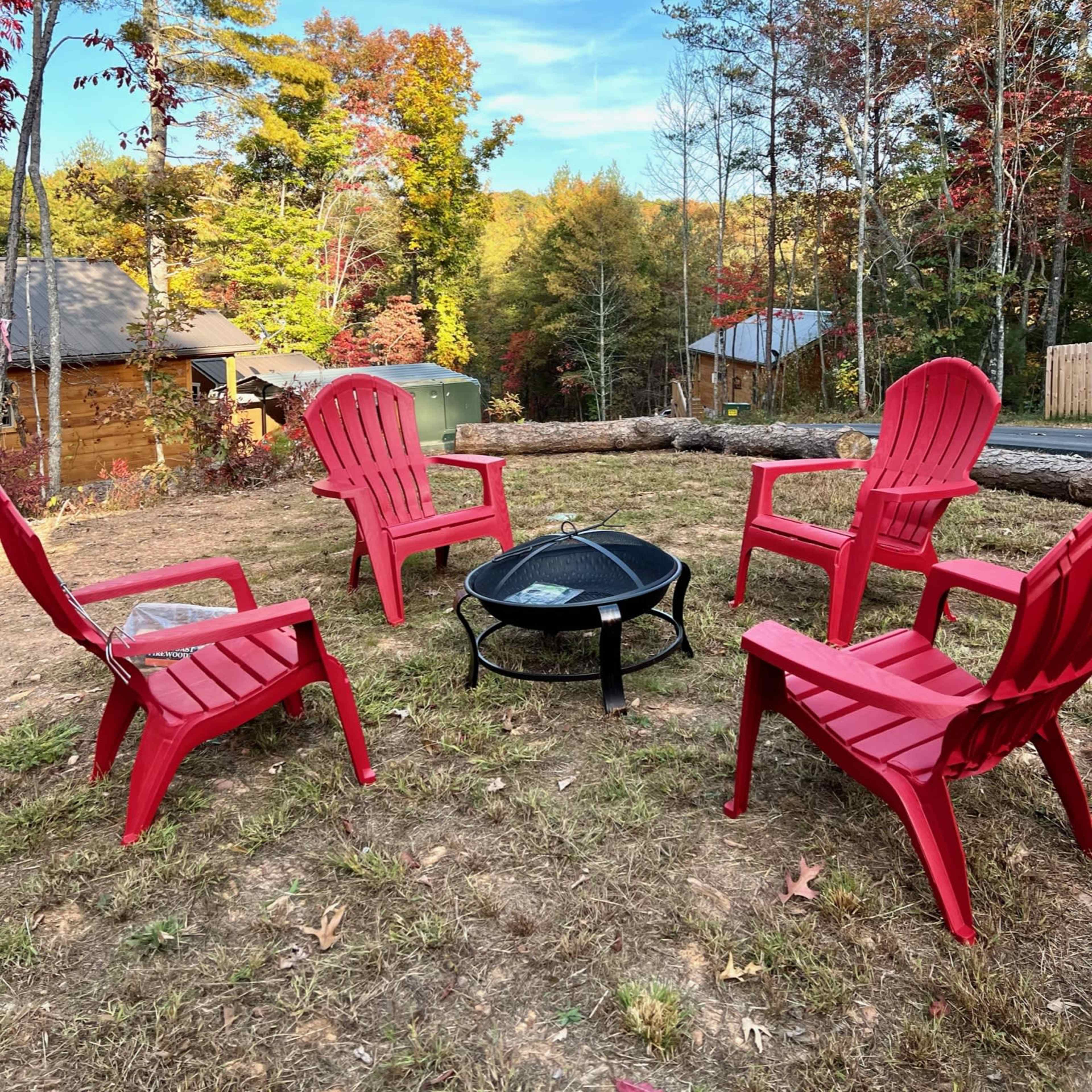 A circular arrangement of red chairs surrounds a black fire pit on a grassy area, with trees displaying autumn colors in the background.