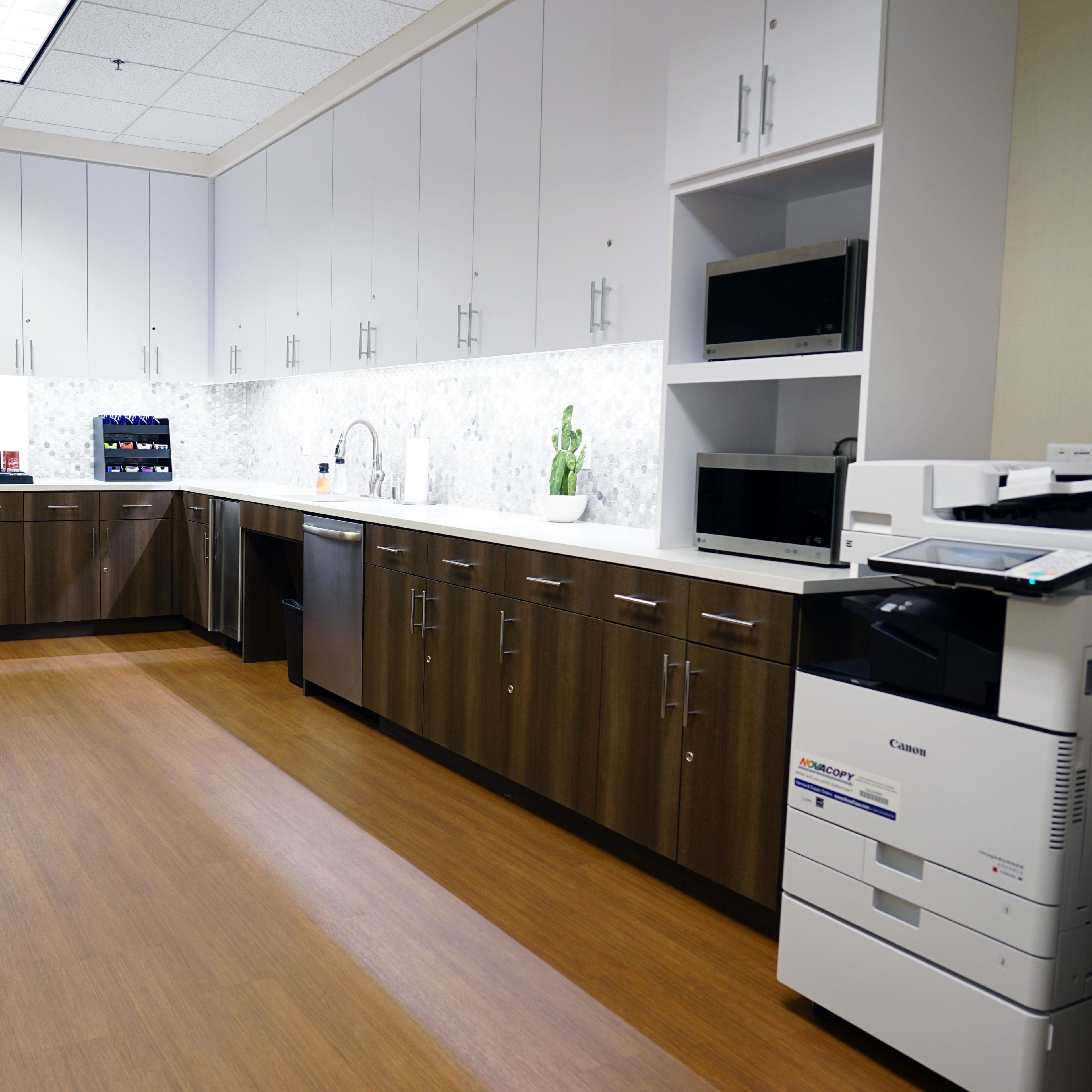 A modern break room with wooden cabinets, a sink, microwaves, a coffee machine, and a printer positioned against a wall.