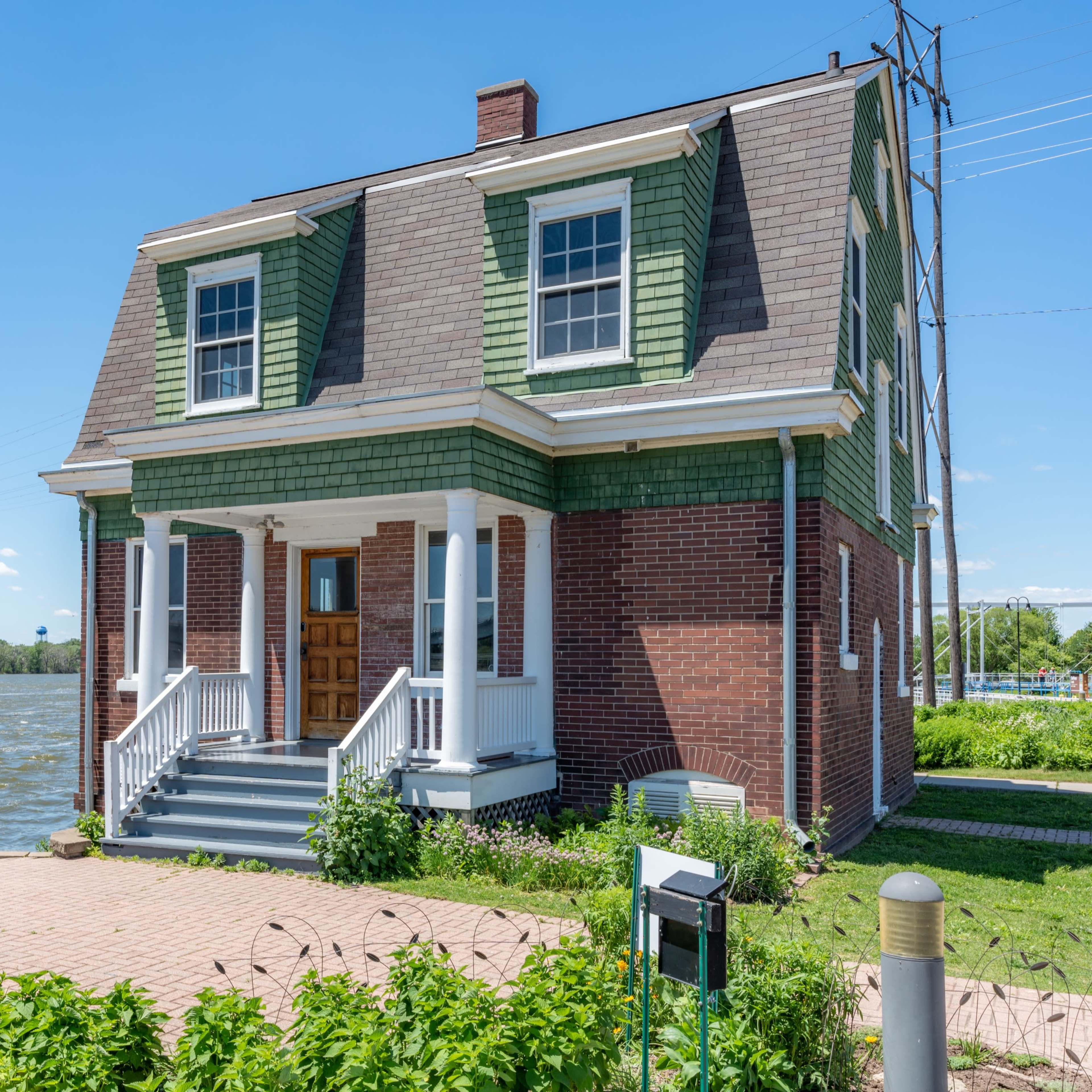 A two-story house with a green and brick exterior is situated near a waterfront, with power lines visible in the background.