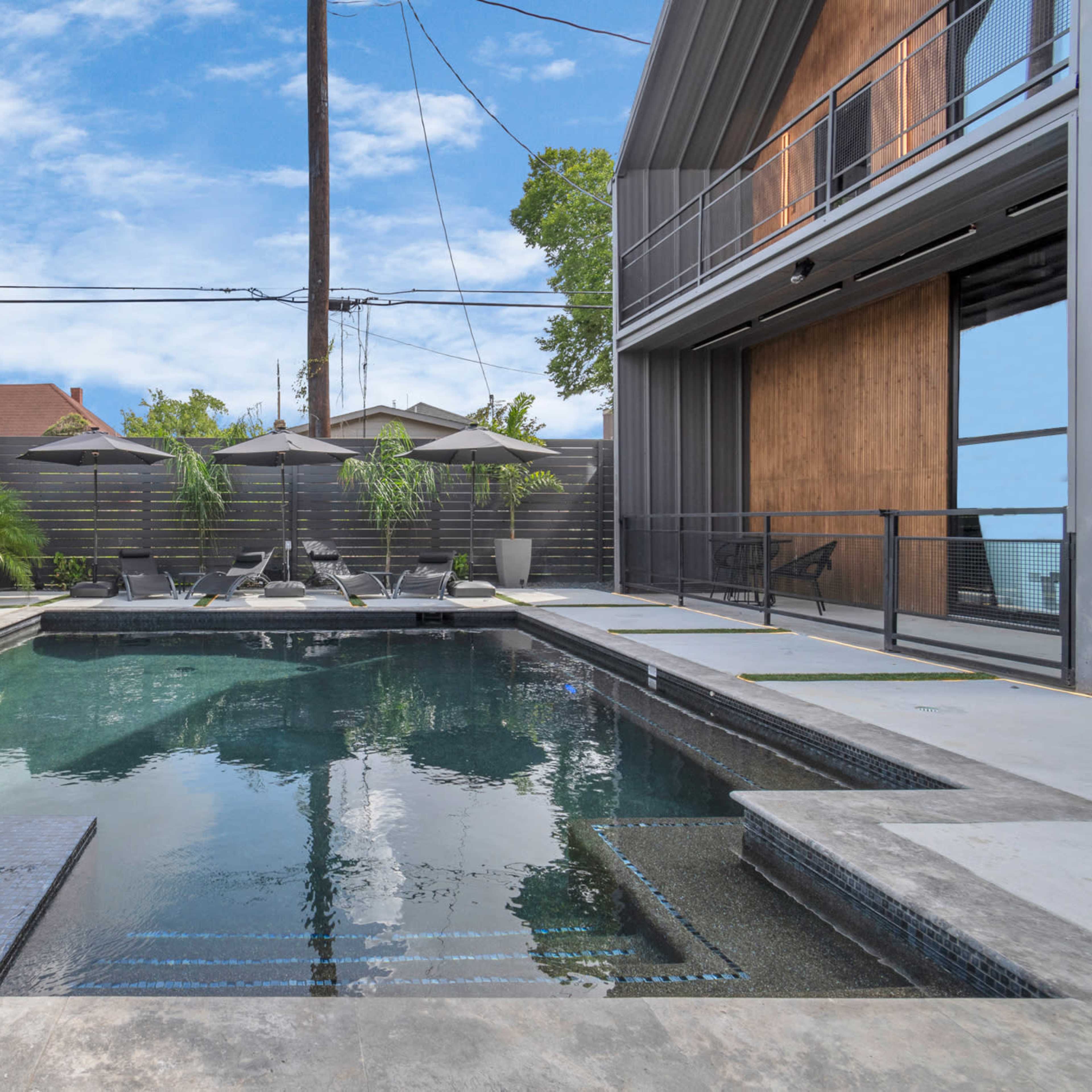A modern swimming pool surrounded by potted plants and lounge chairs, with a two-story building in the background.