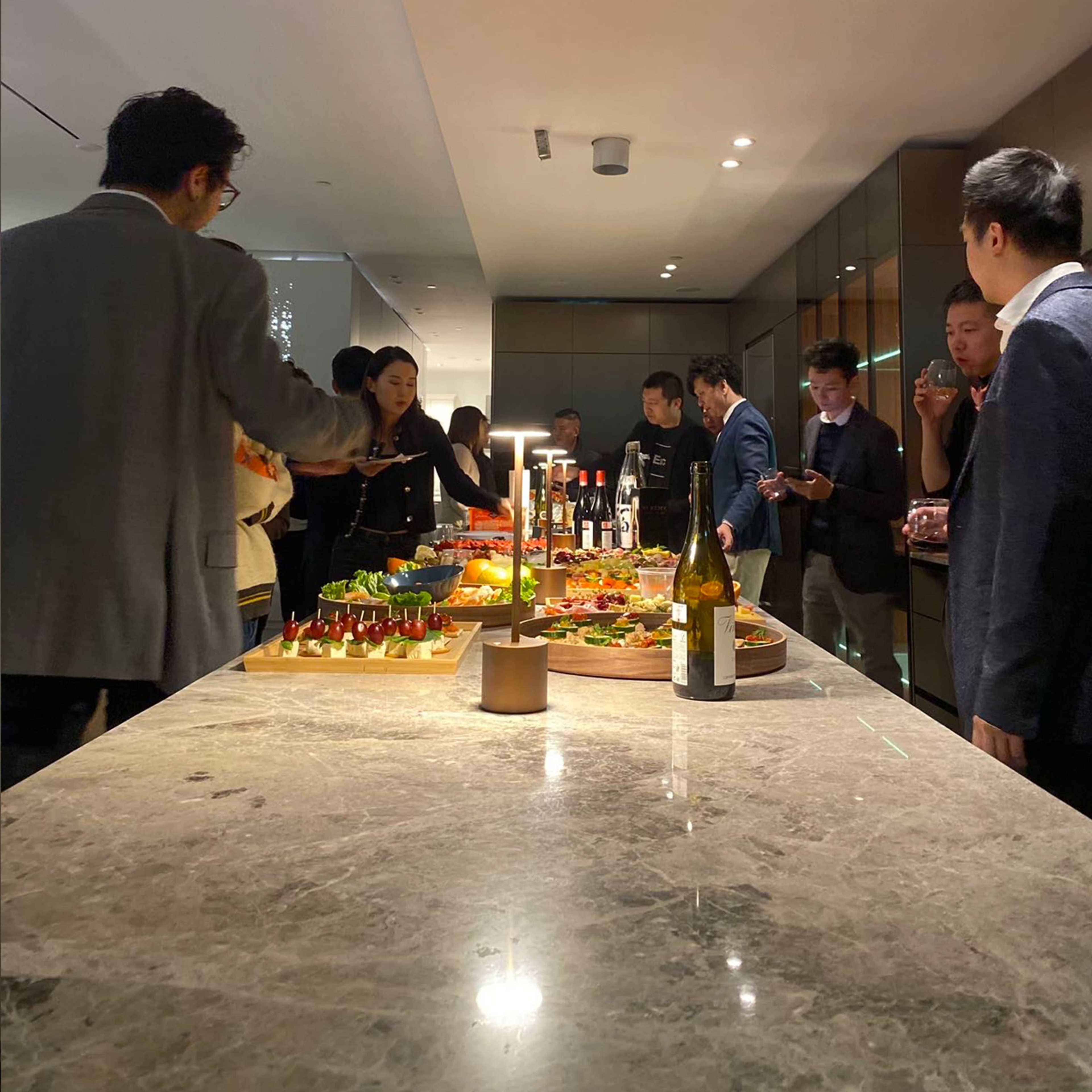 A group of people gathers around a marble countertop filled with various food platters and drinks in a modern lounge setting.