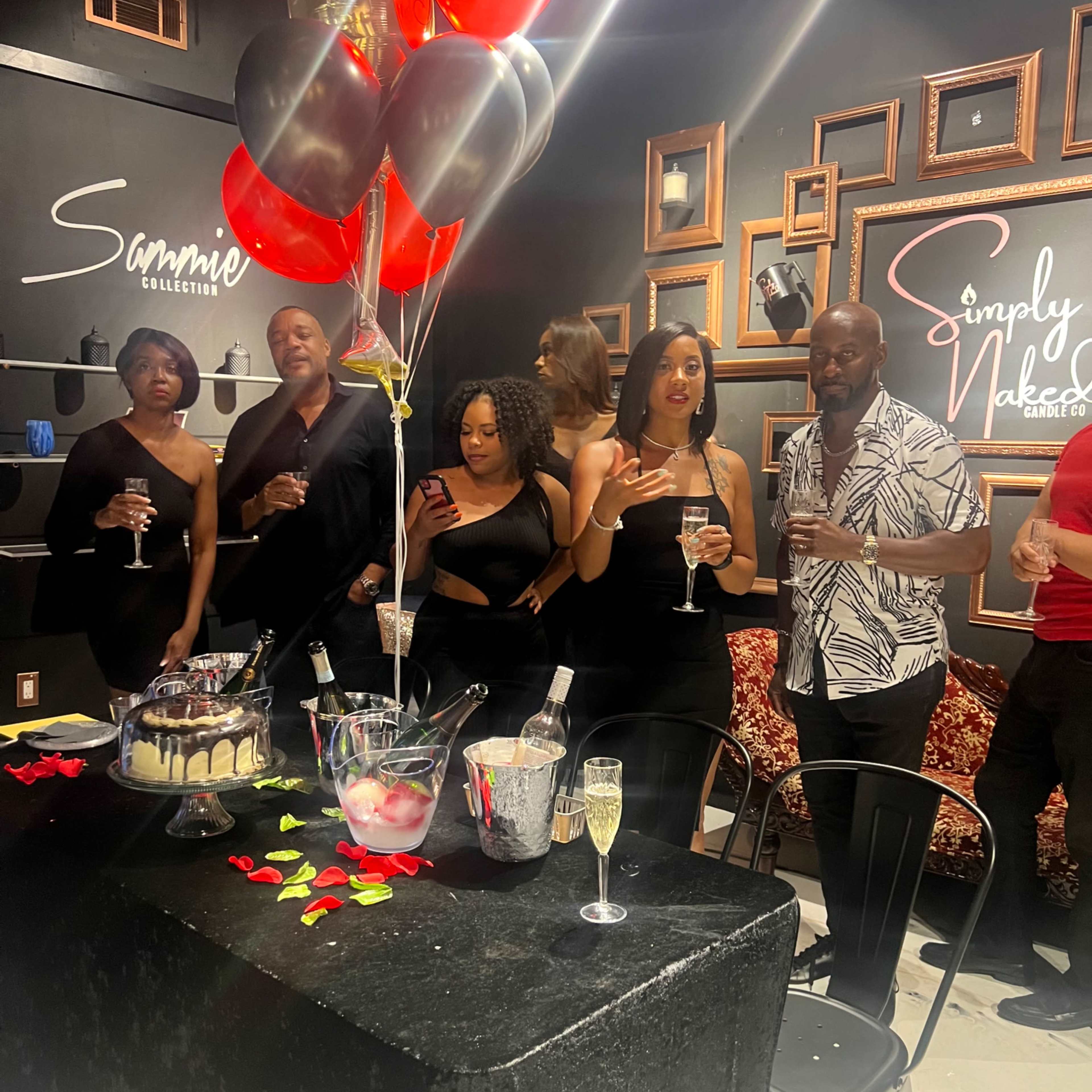 A group of people stands around a festively decorated table with drinks and balloons at a social gathering.