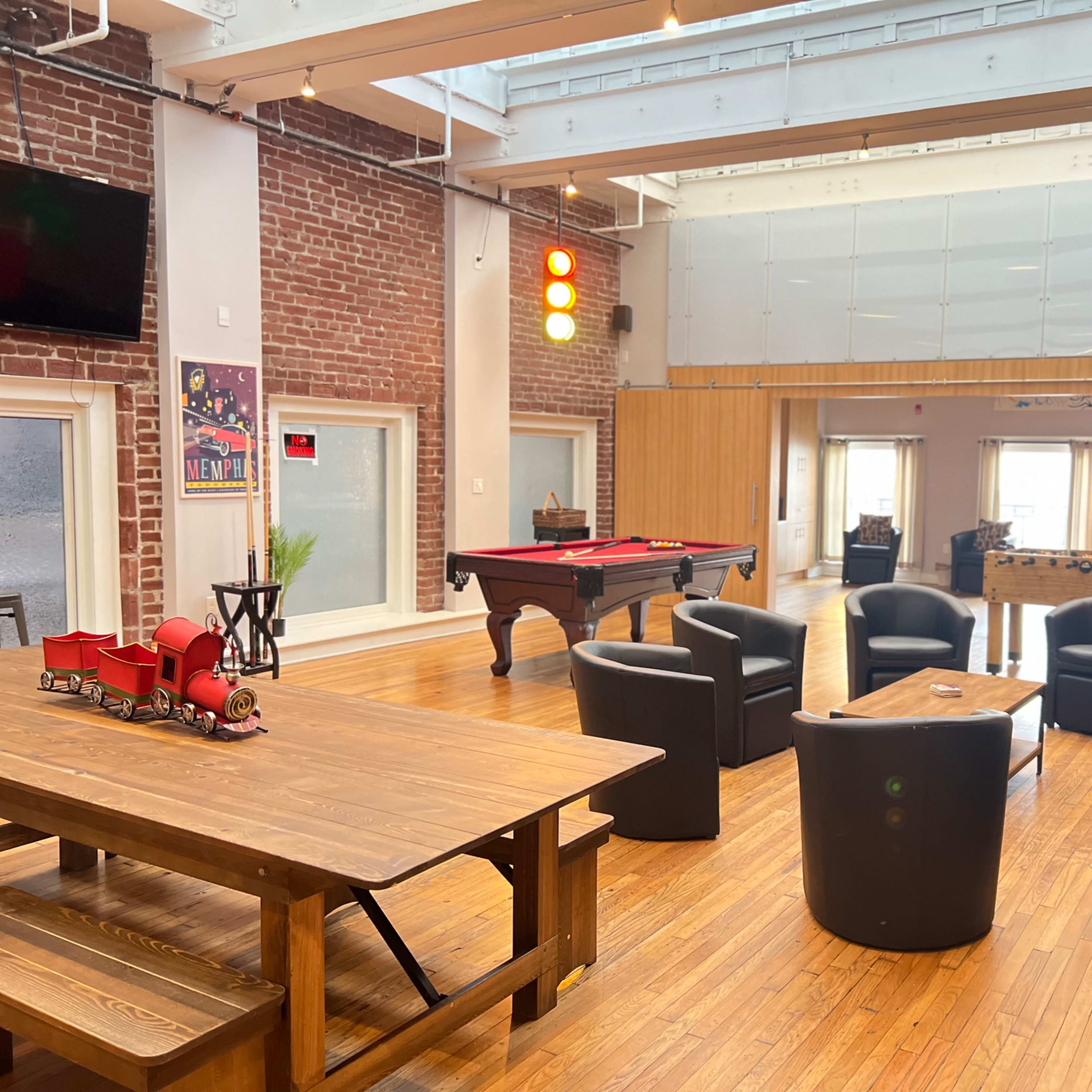 A spacious room with wooden flooring, featuring a pool table, foosball table, and seating areas with black chairs and a couch, alongside a large wooden dining table and a barrel.