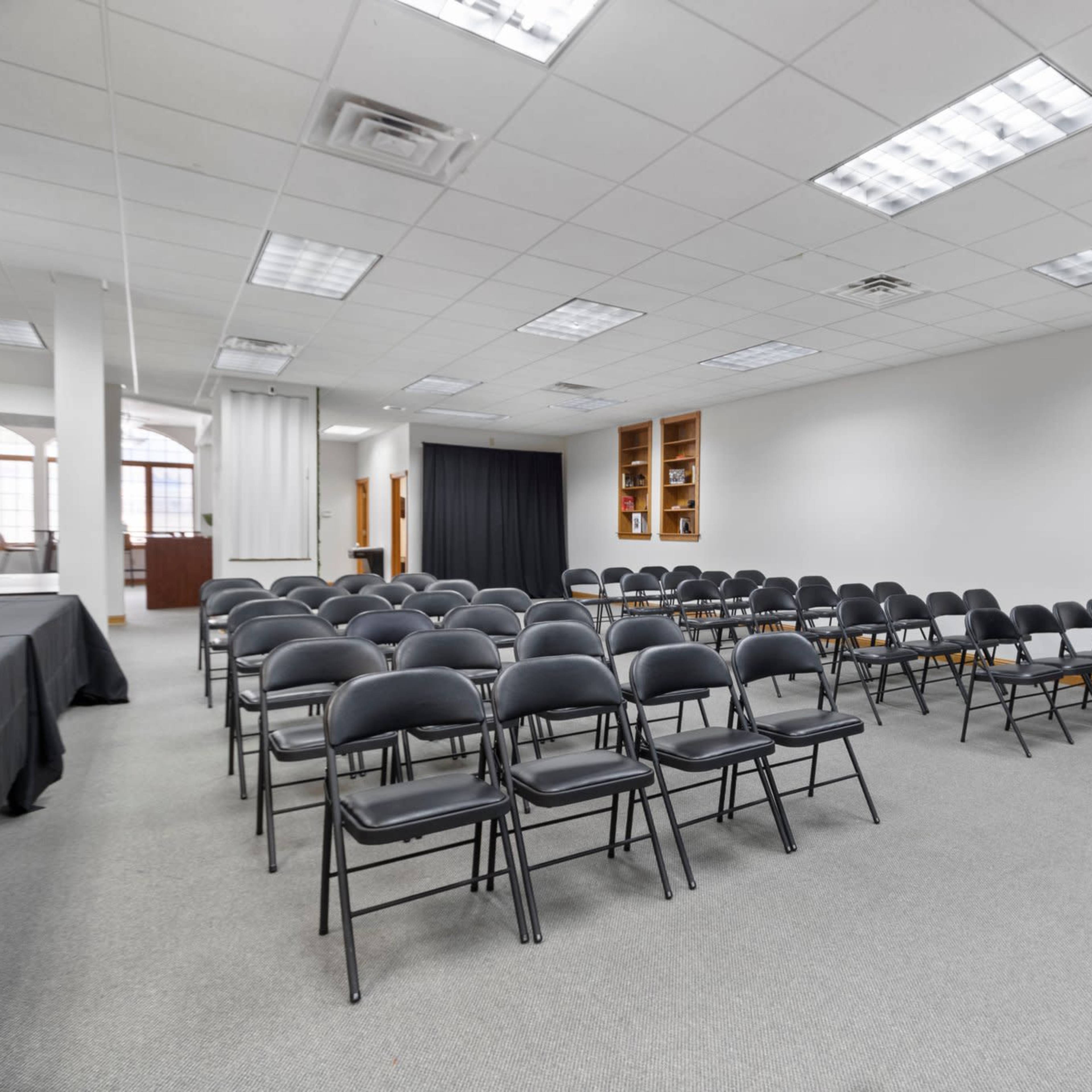 A meeting room set up with rows of black folding chairs facing a black wall, with a few tables along one side.