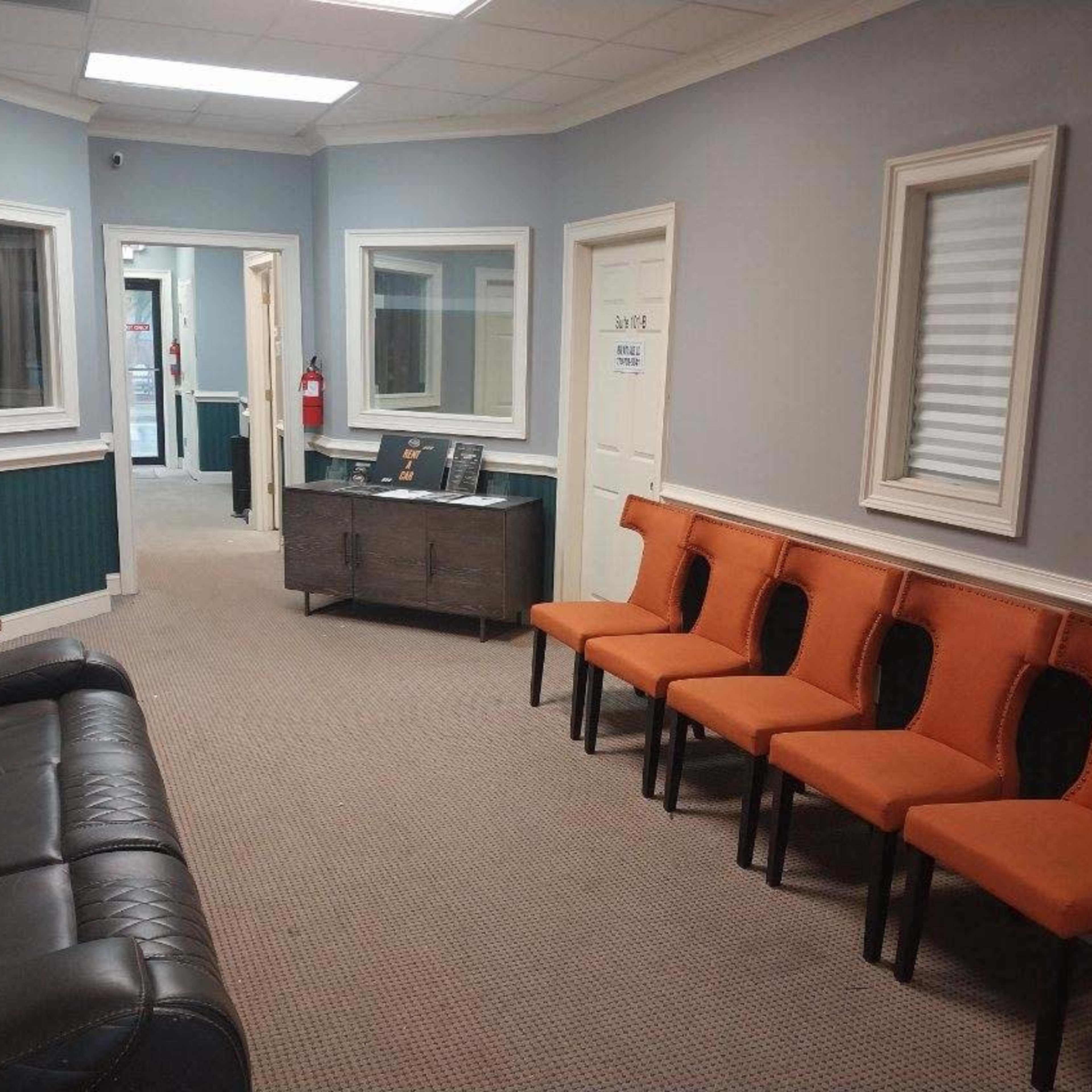A waiting room with a black leather couch and a row of orange chairs against the wall, framed by light gray walls and two doors leading to other rooms.