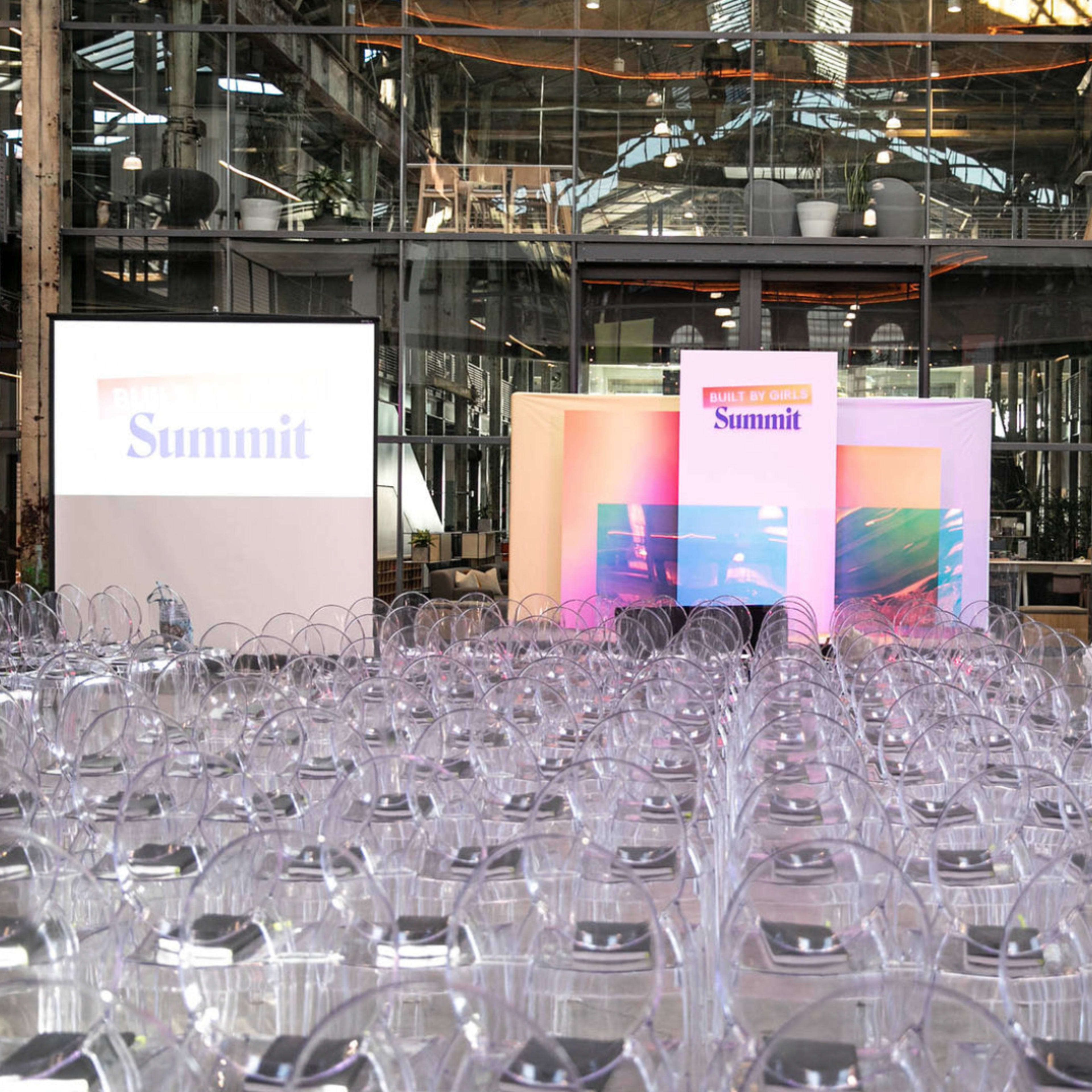 A large indoor event space filled with transparent chairs arranged in rows facing a stage with colorful banners labeled "Summit."
