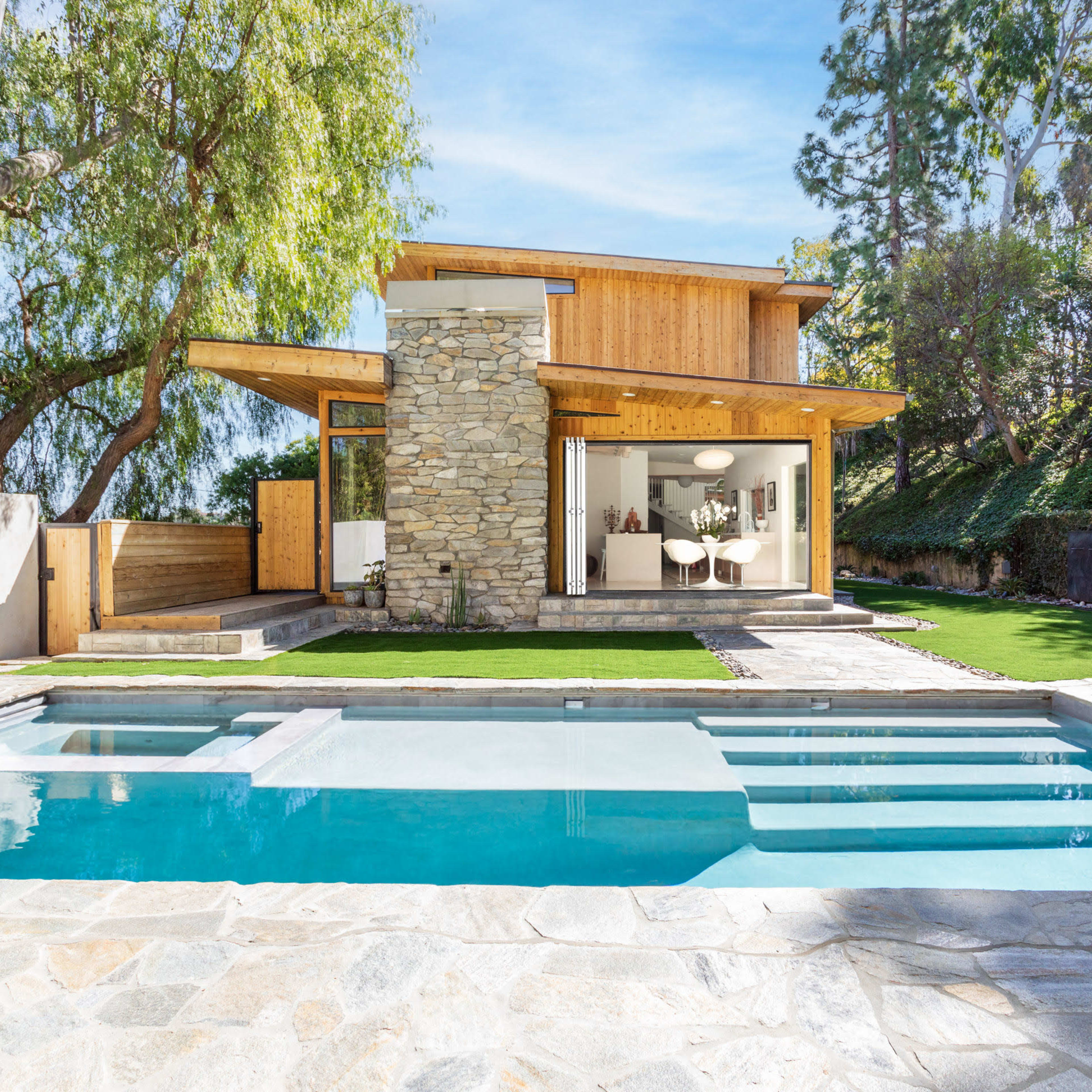 A modern house with a stone facade features large windows and a pool in the foreground, surrounded by landscaped greenery.