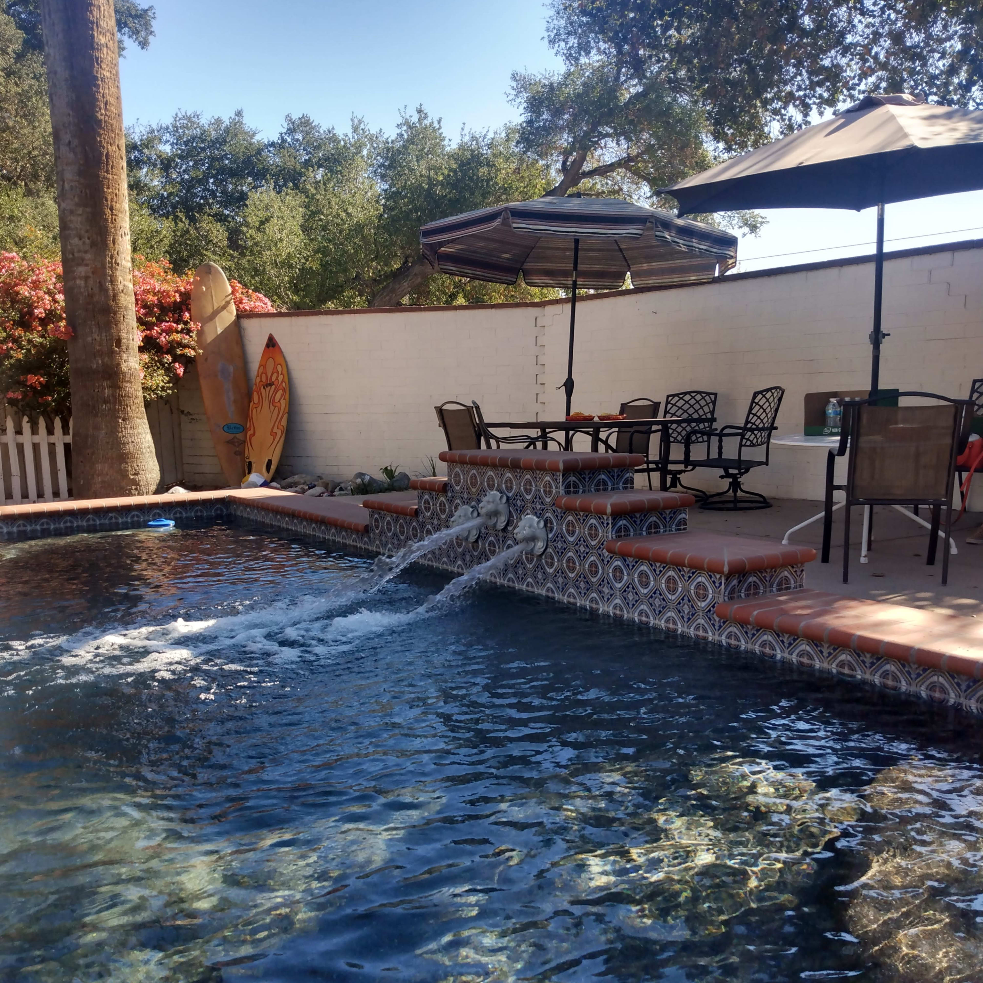 A swimming pool with a fountain feature is surrounded by patio furniture and shaded by umbrellas, with a surfboard resting against a wall in the background.
