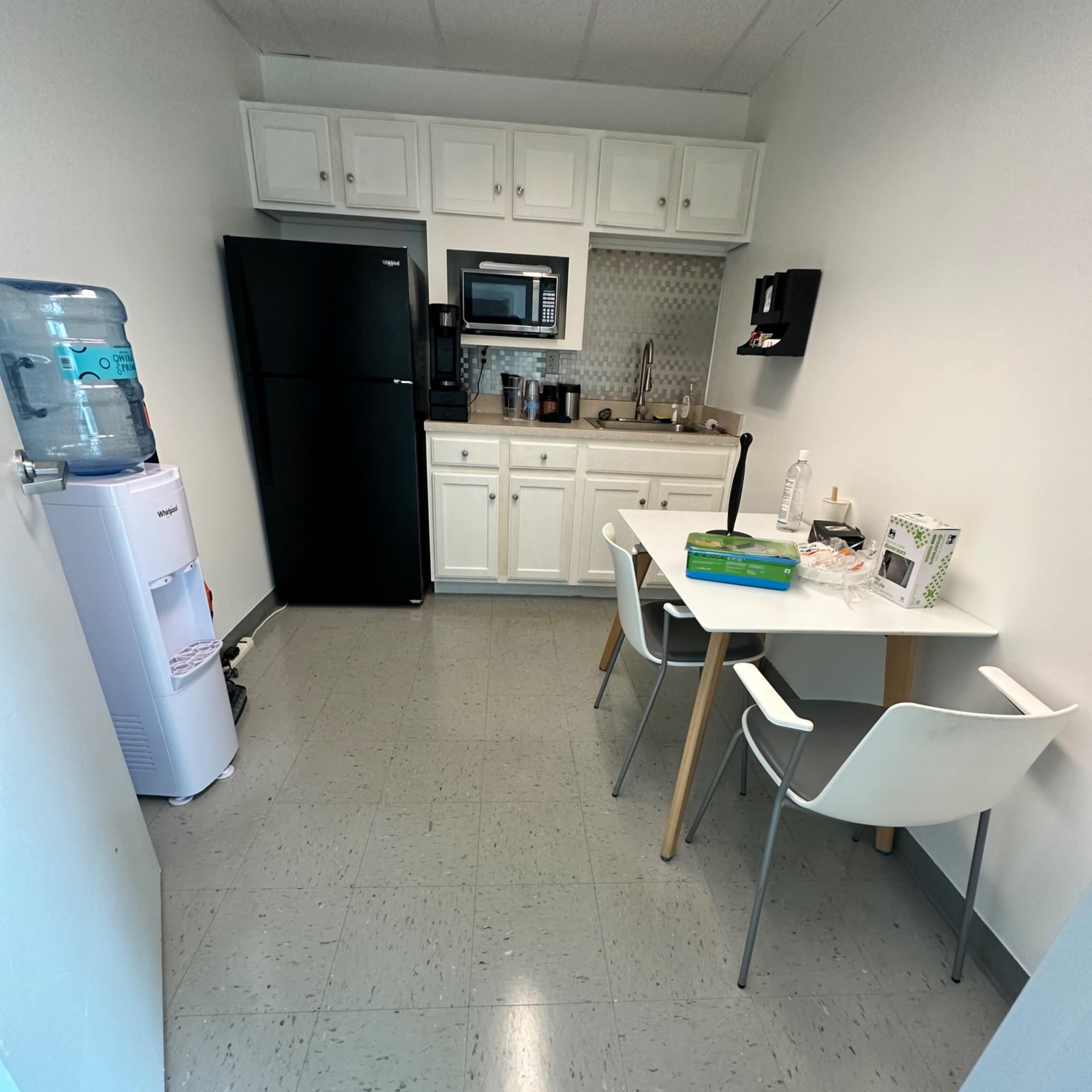 A small kitchen area featuring a black refrigerator, a microwave, a table with two chairs, and a water cooler.
