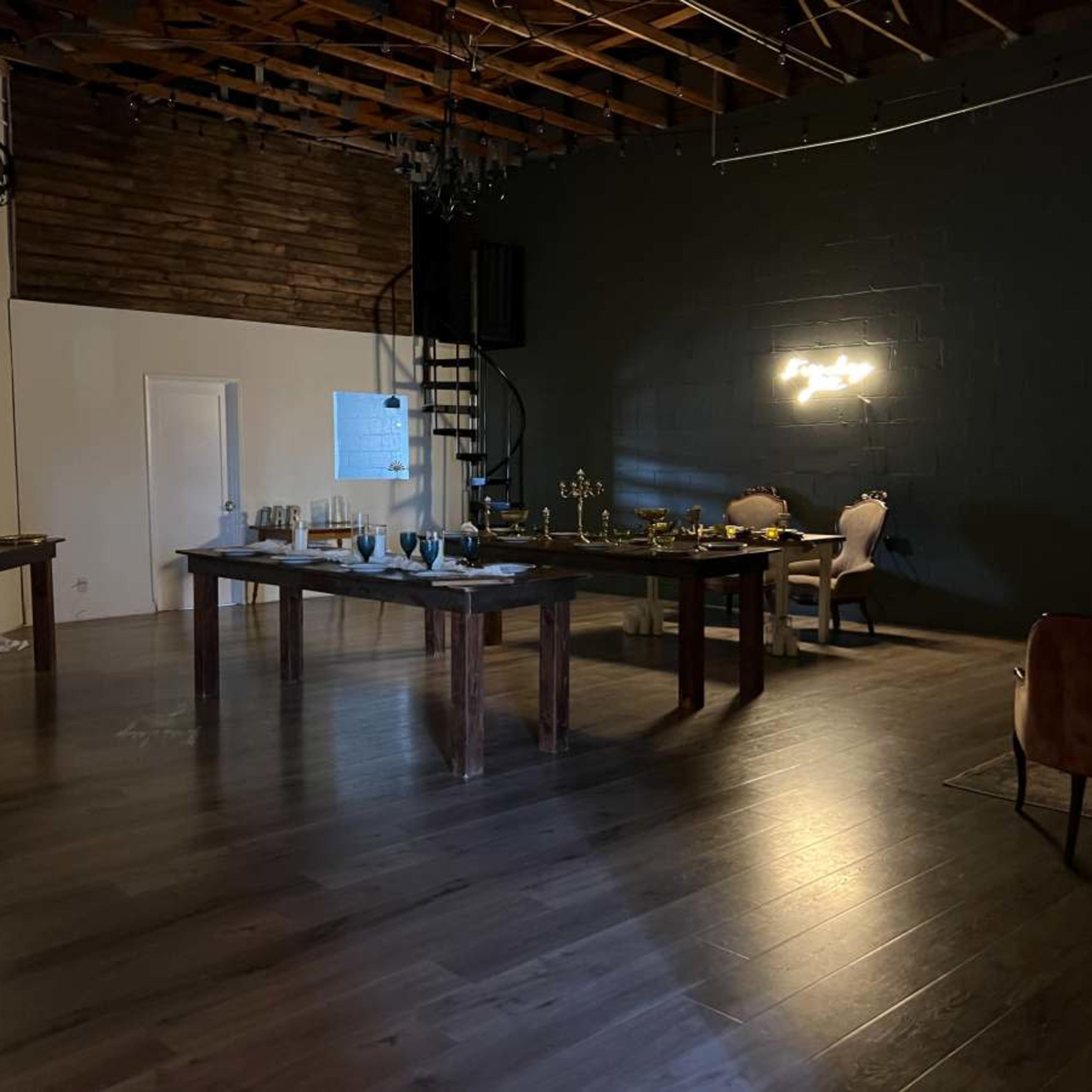 A spacious indoor venue with wooden floors, round tables set for dining, and a spiral staircase in the background.