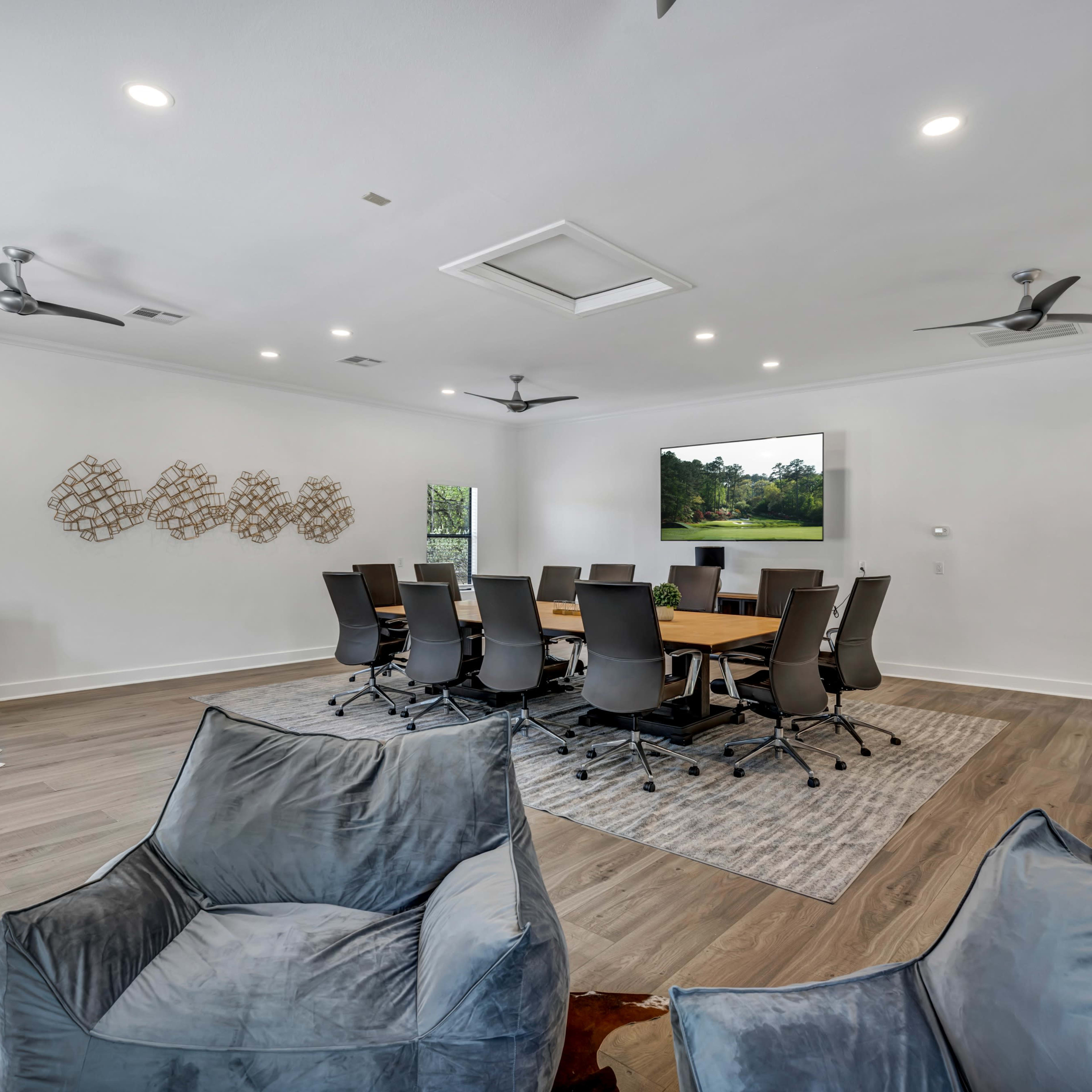 A modern conference room features a large table surrounded by ergonomic chairs, with a large screen on one wall and casual seating in the foreground.