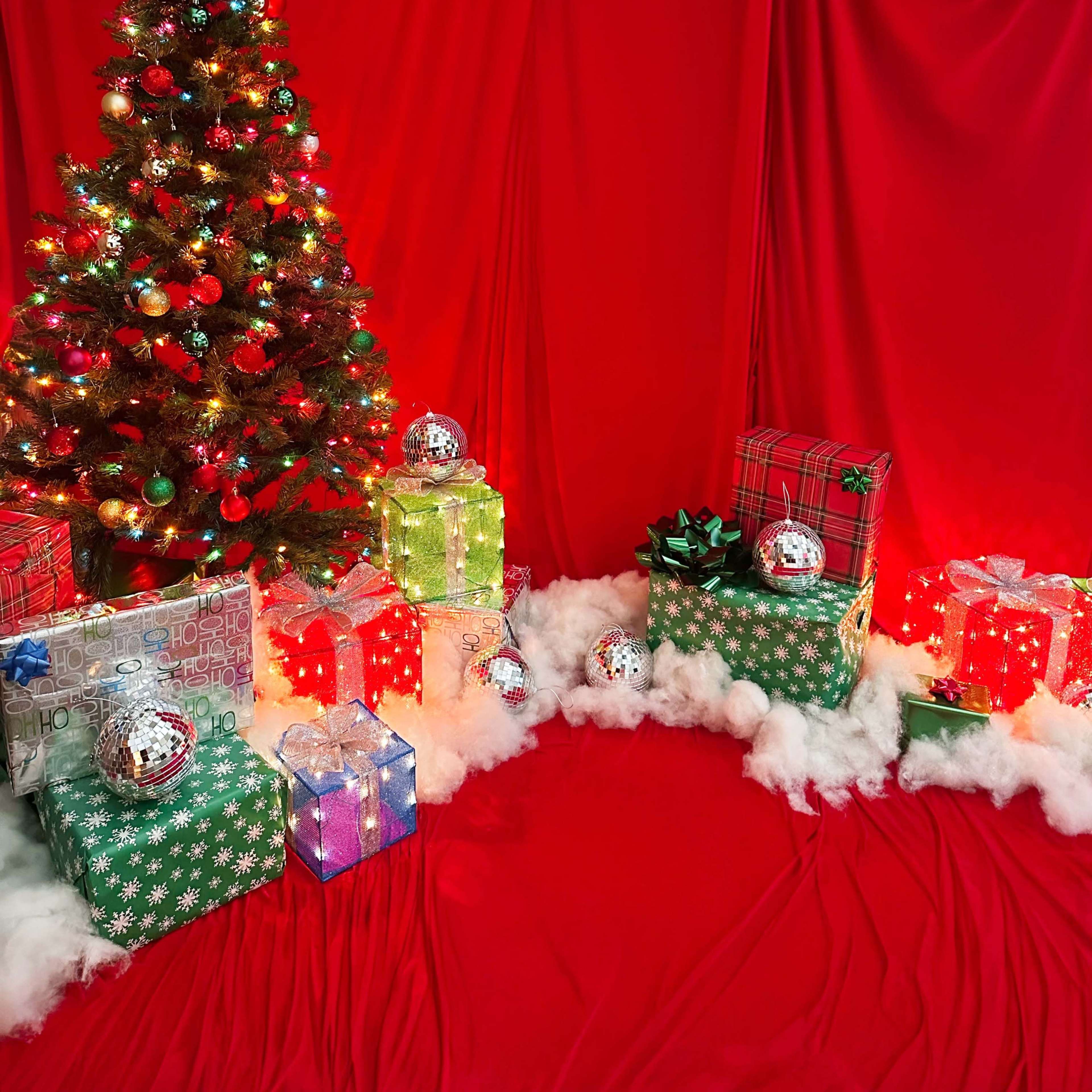 The scene features a Christmas tree adorned with colorful lights and ornaments, surrounded by wrapped gifts in various colors on a red backdrop.