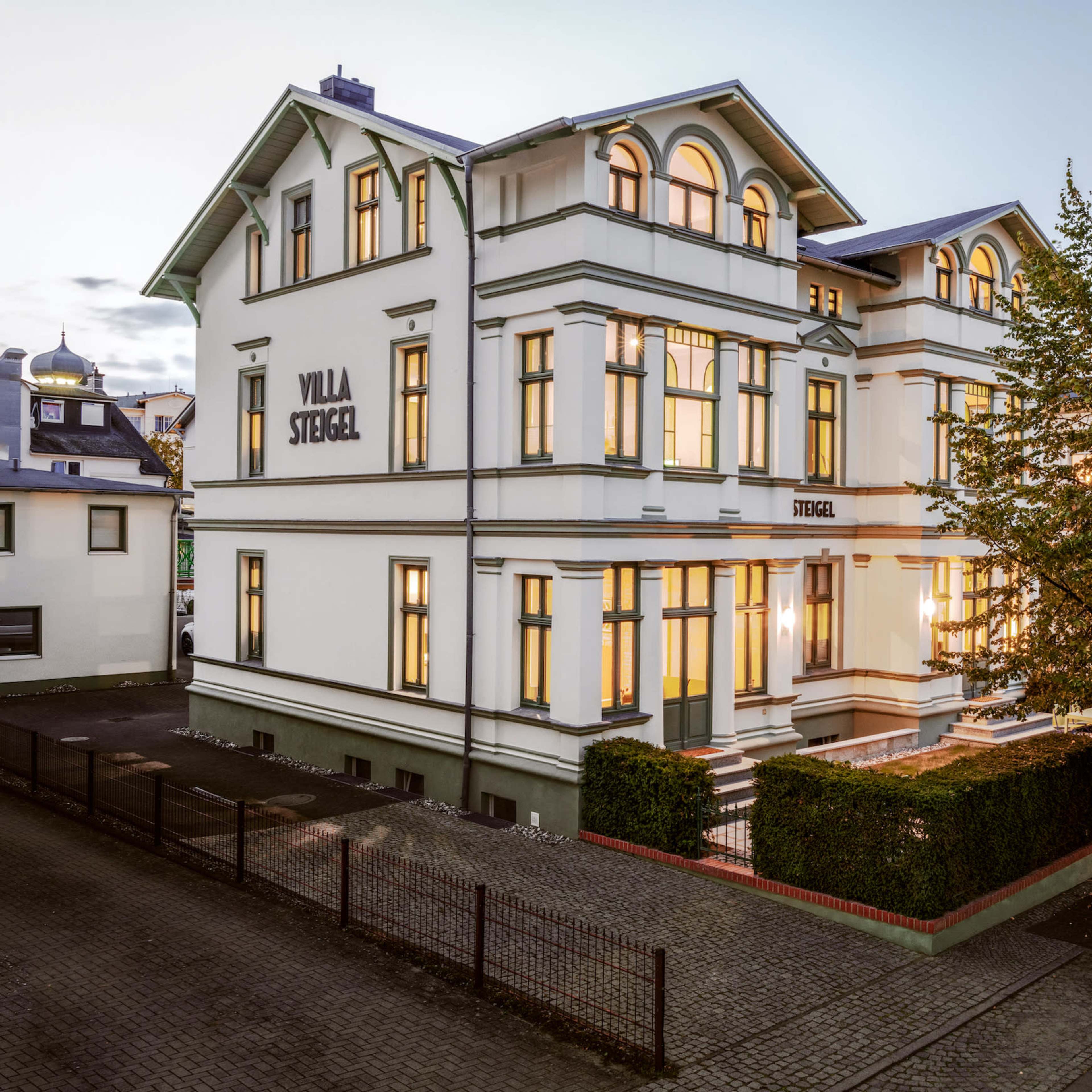 A large, white, historic villa with multiple windows and a manicured garden, situated on a street corner at dusk.