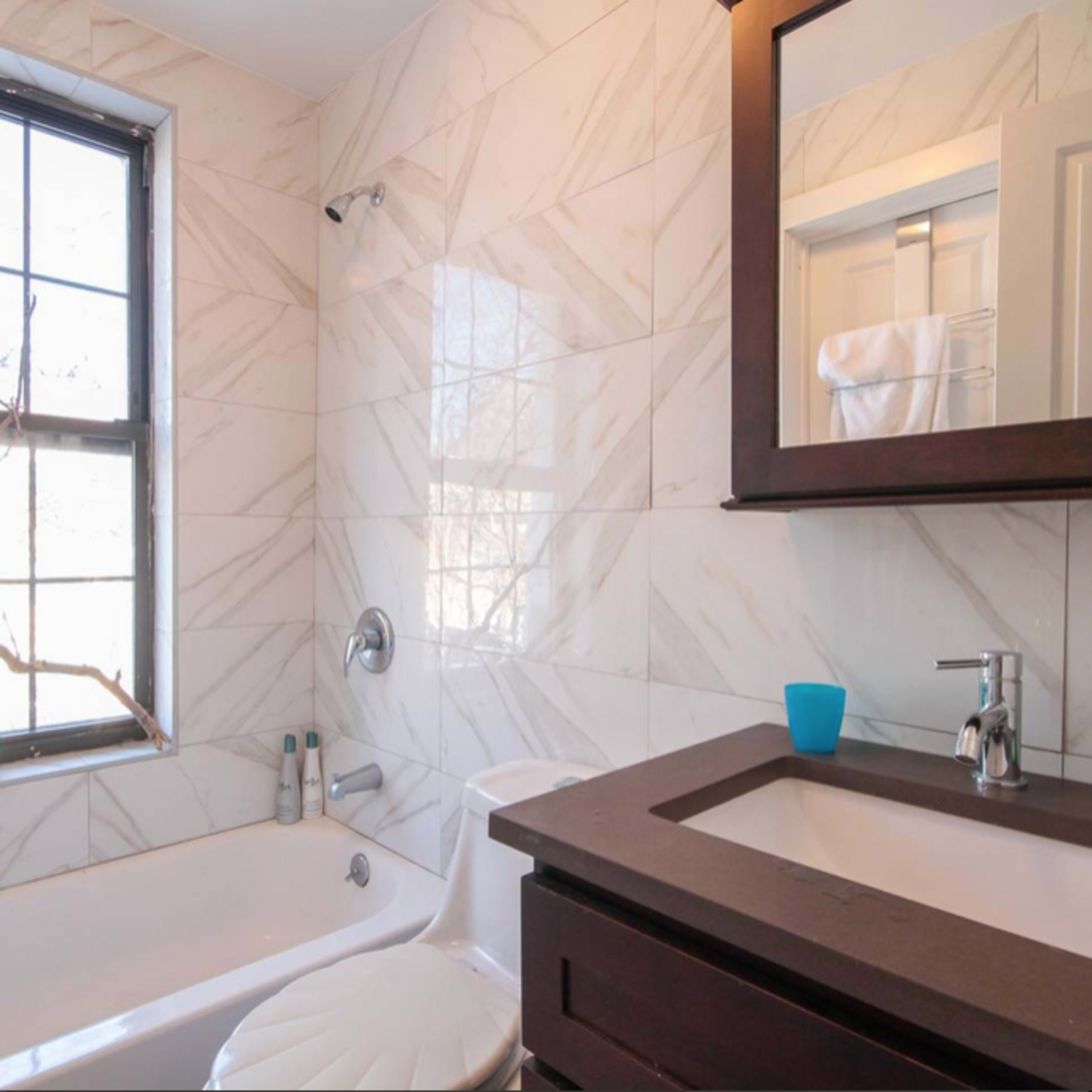 The bathroom features a bathtub, a wall-mounted showerhead, a sink with a dark countertop, and large windows allowing natural light to enter.