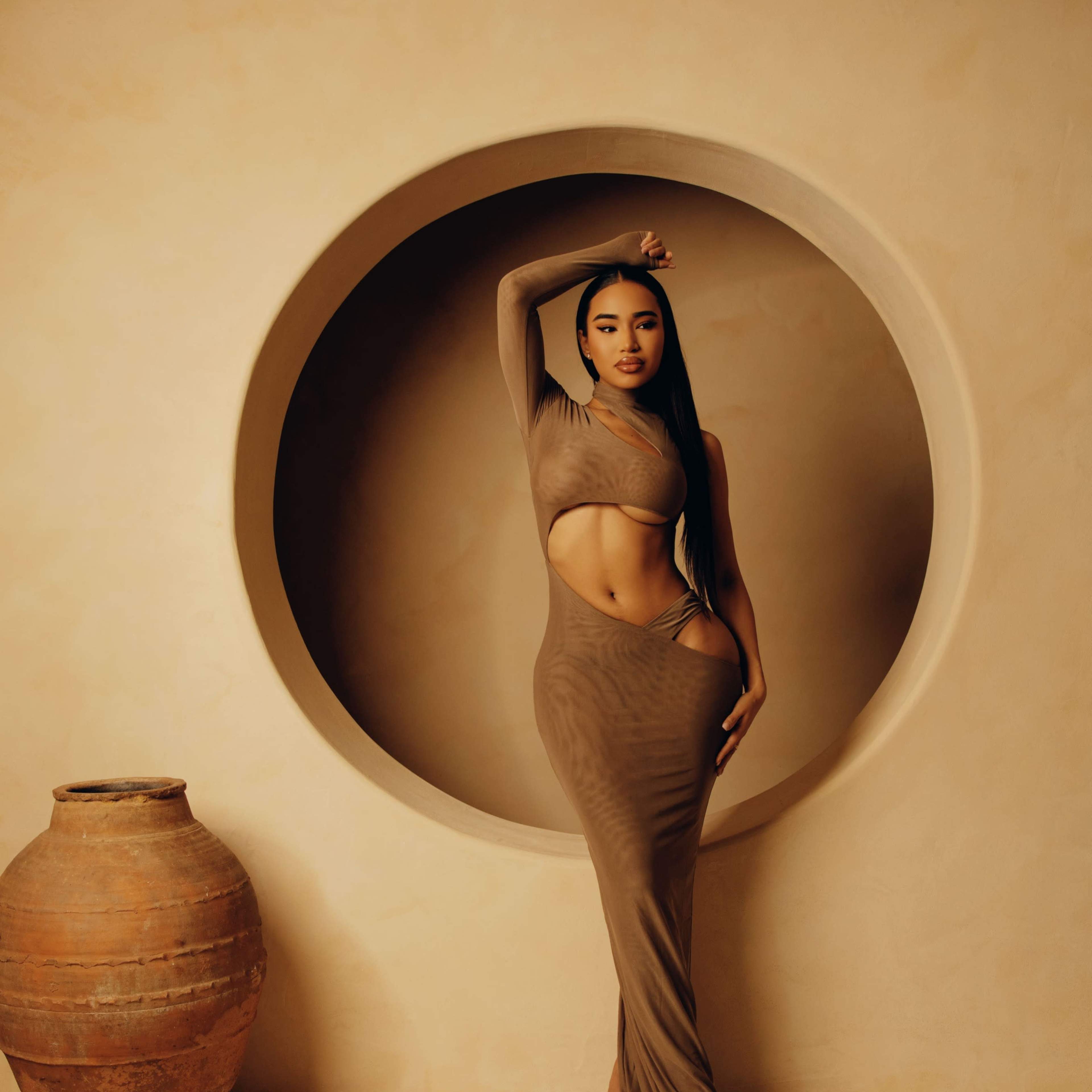 A woman in a fitted brown dress poses beside a large round recess in a textured beige wall, with a clay pot nearby.