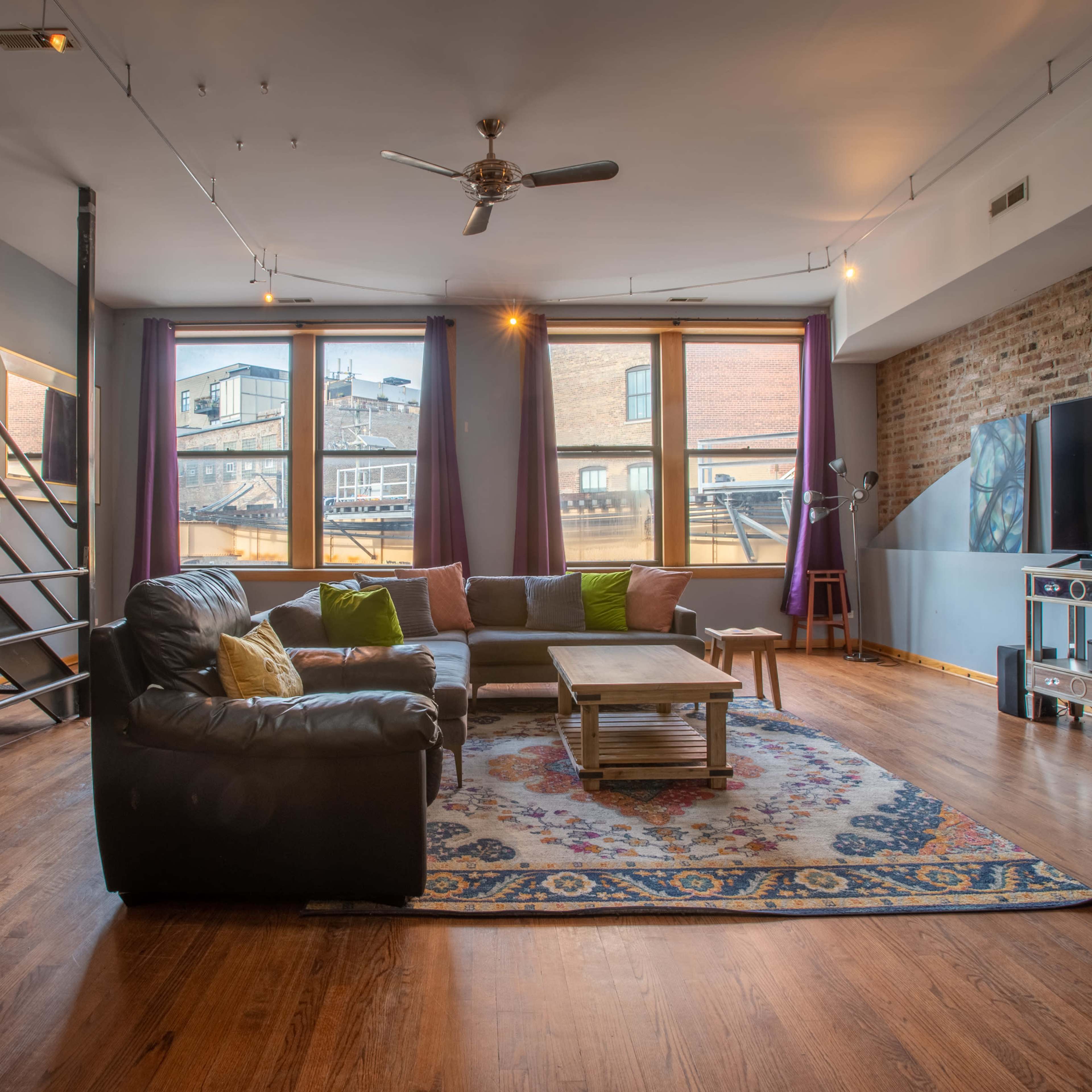 A spacious living room featuring a black leather sectional sofa, a wooden coffee table on a patterned rug, and large windows with views of a cityscape.