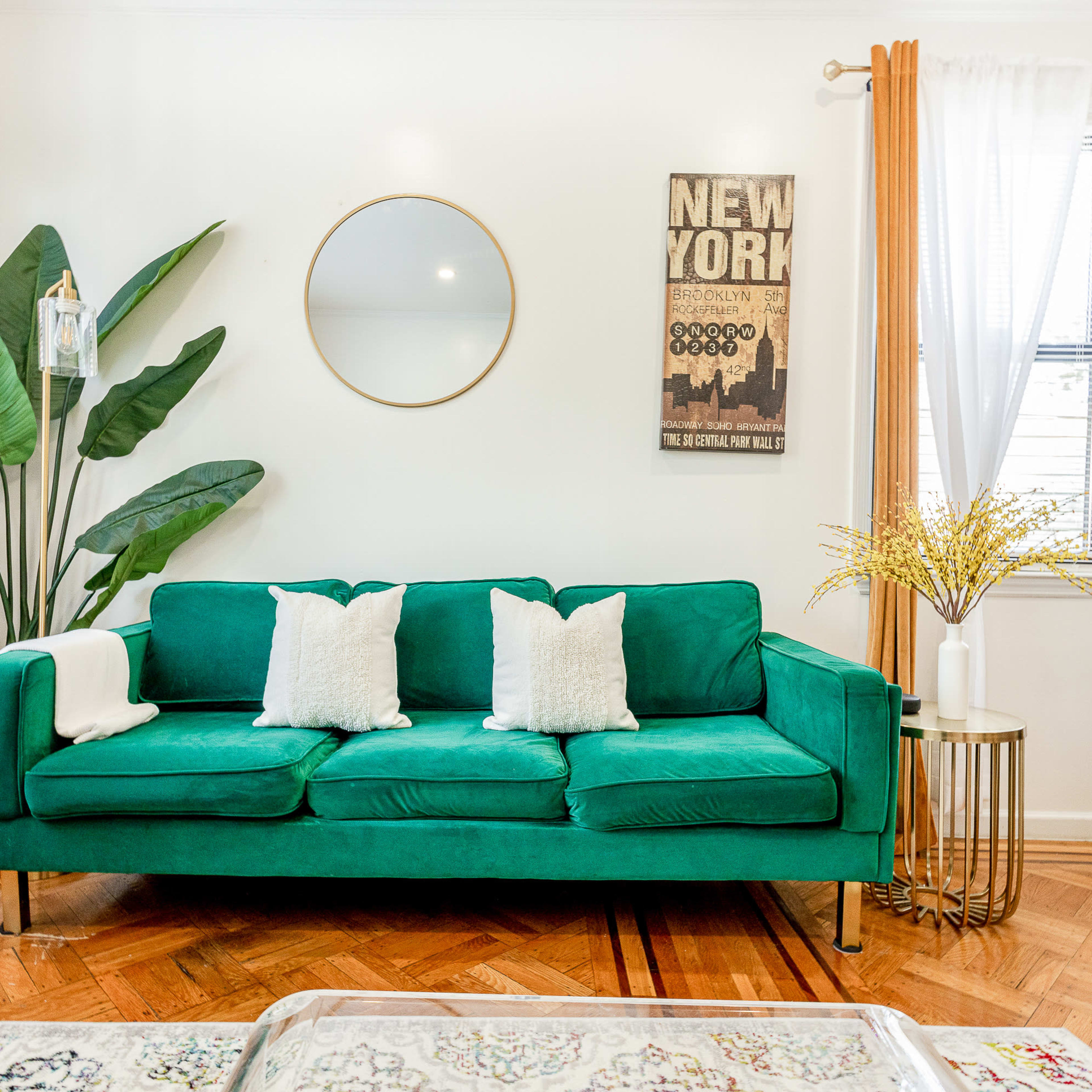 A green sofa with white pillows is positioned in a brightly lit room featuring plants, a round mirror, and decorative curtains.