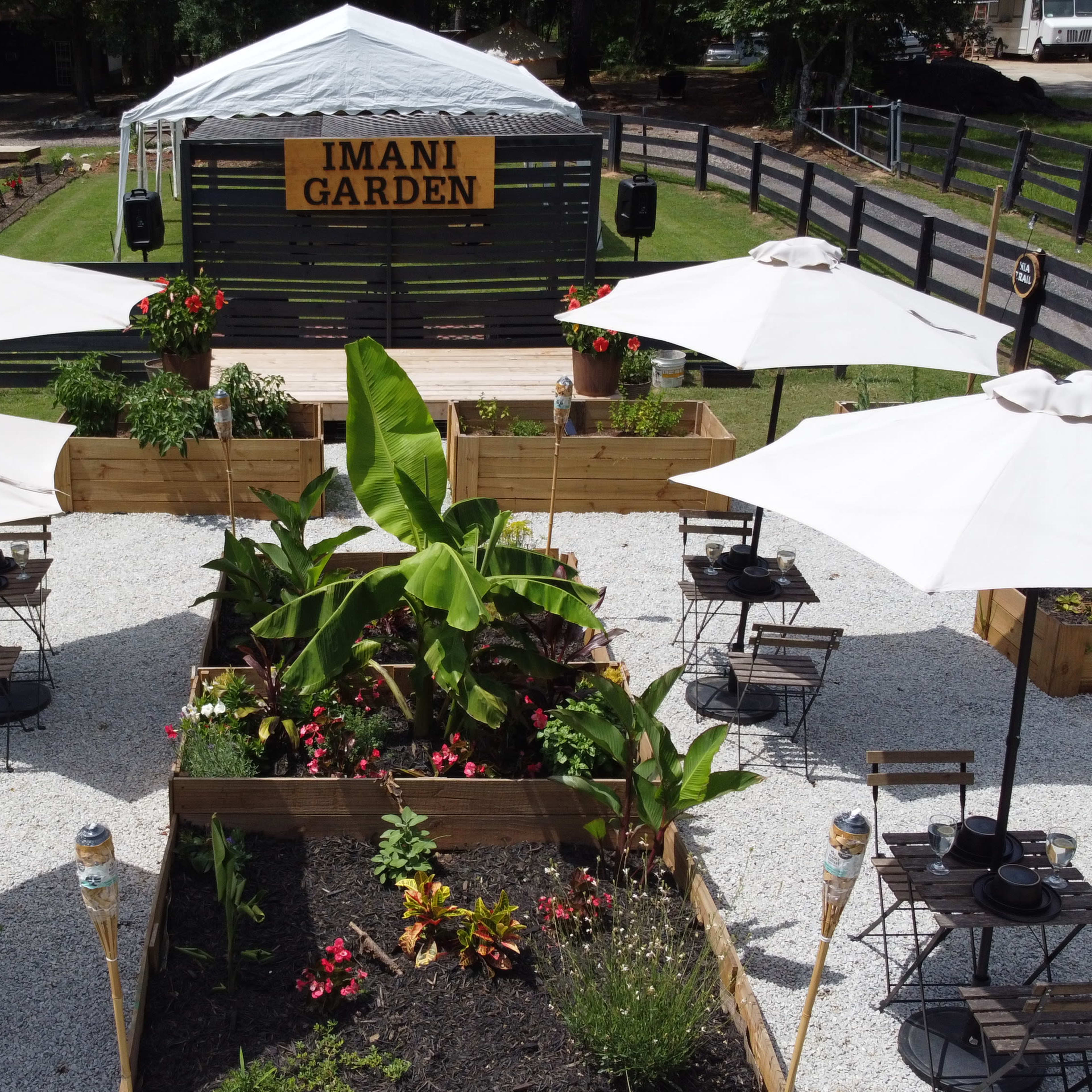 A garden space called "IMANI GARDEN," featuring wooden planters, tables with chairs under white umbrellas, and decorative plants arranged in a gravel layout.