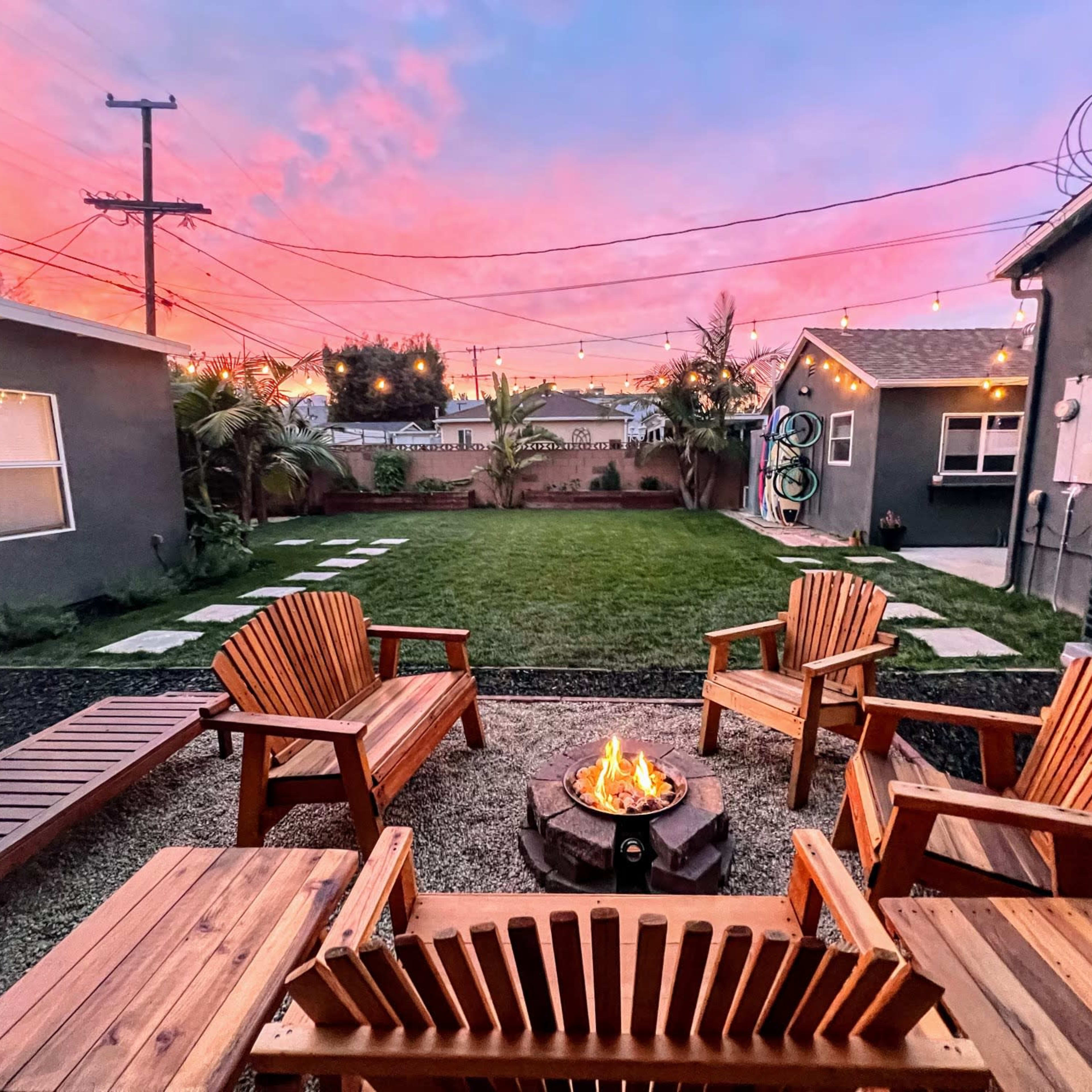 A cozy outdoor space features four wooden chairs surrounding a fire pit, with a well-maintained lawn and colorful sunset skies in the background.