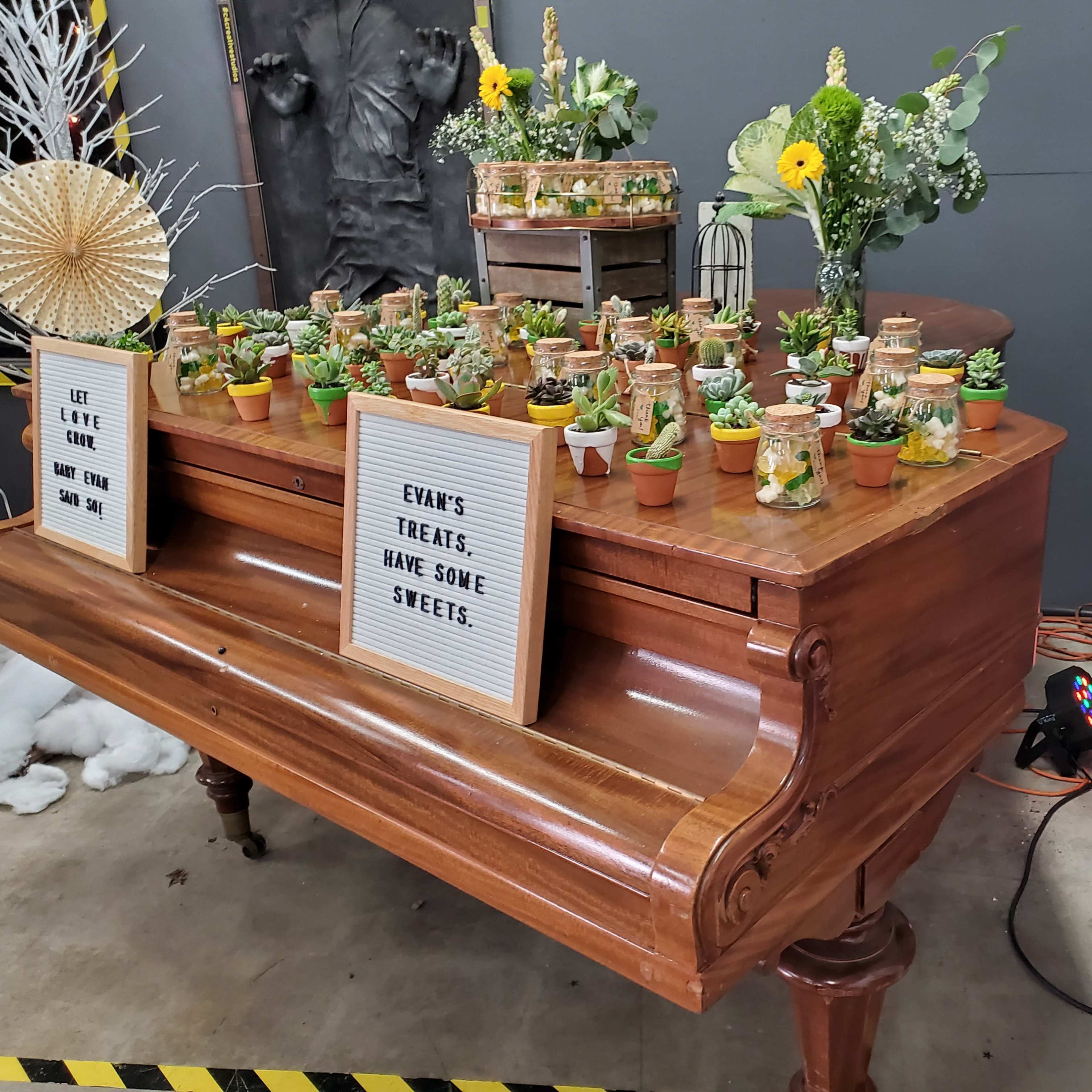 A wooden table displays small potted plants alongside jars of sweets, adorned with decorative signs and flowers in a festive setting.