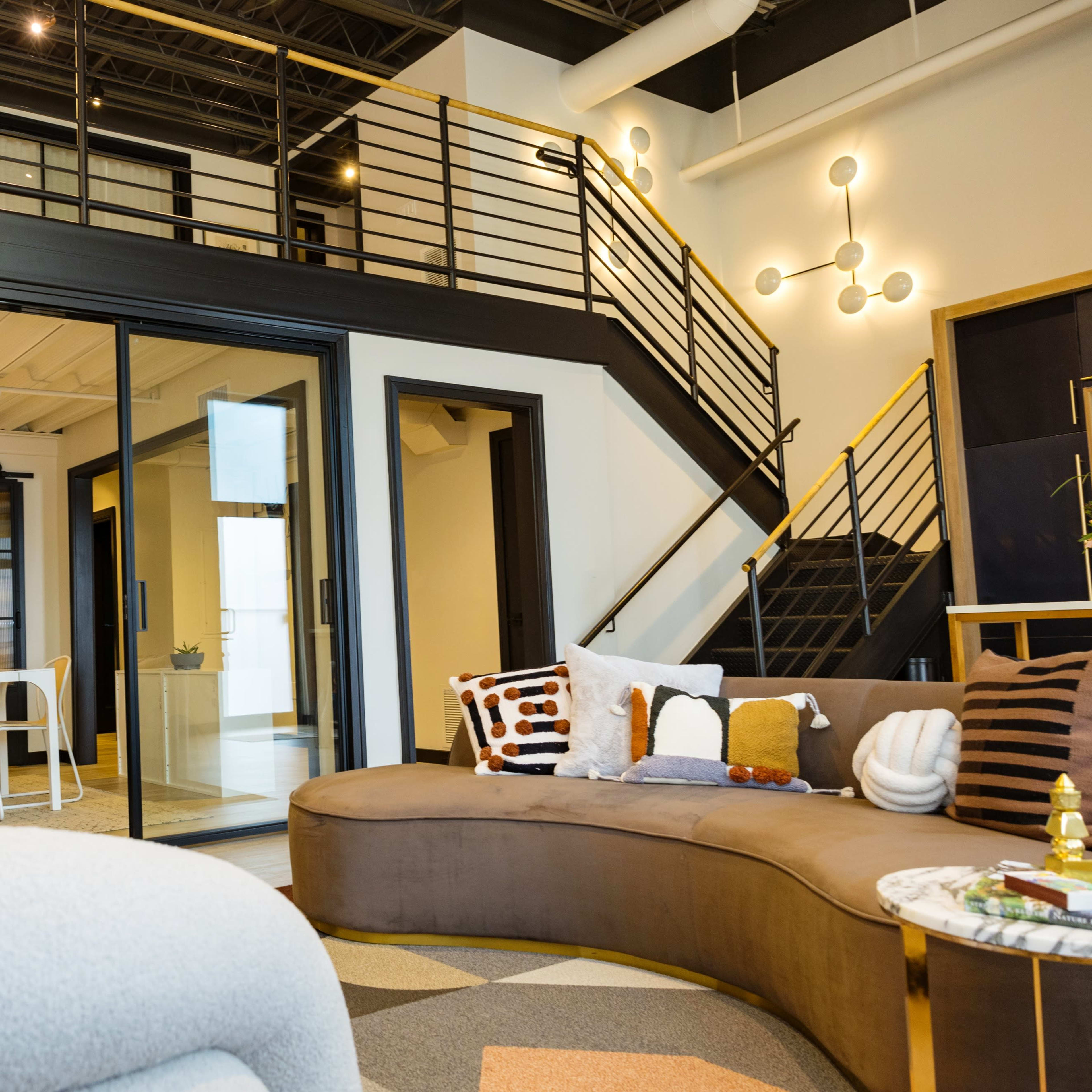 A modern, open-concept interior with a curved sofa, a dining area with white chairs, and a staircase leading to an upper level.