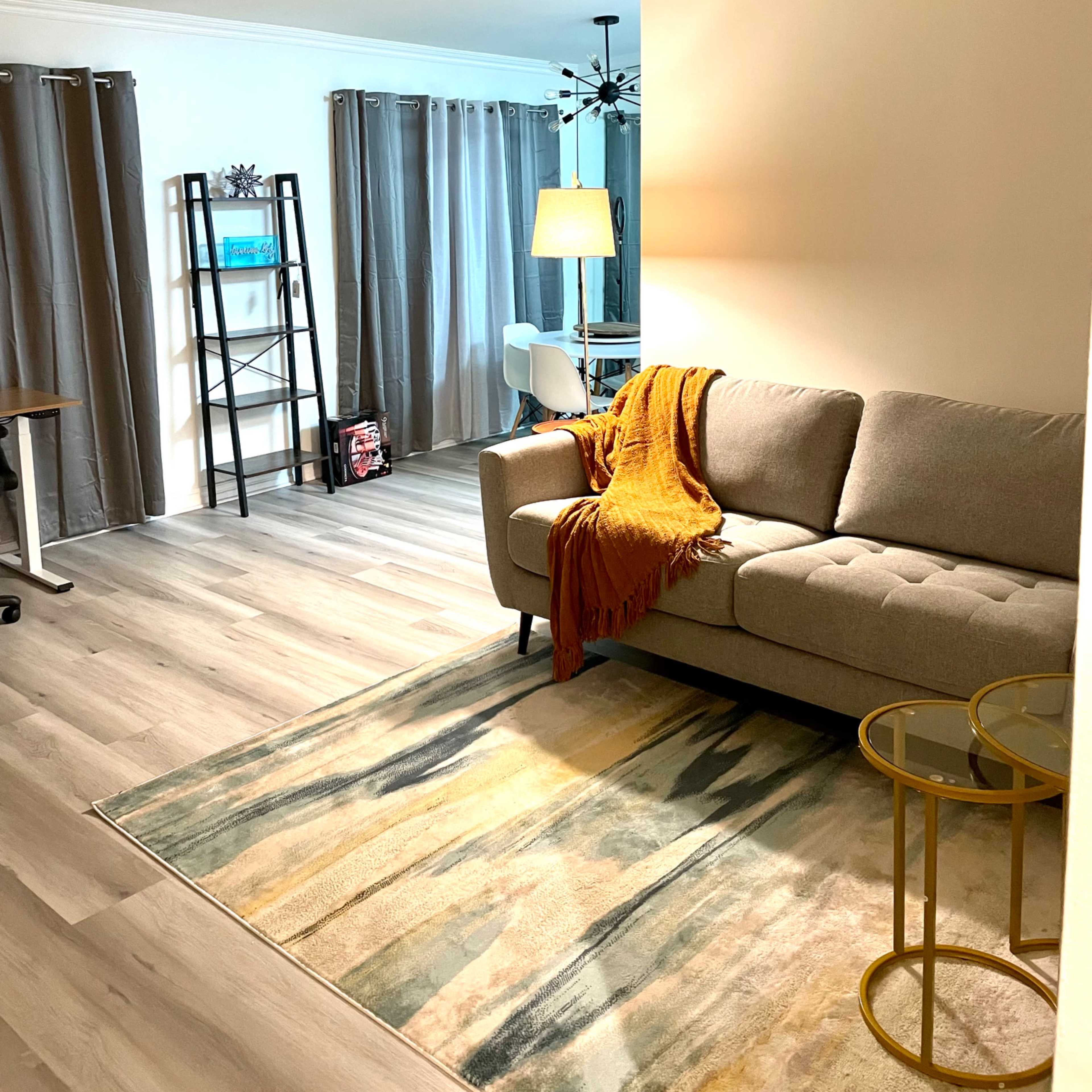 A neatly arranged living room with a gray sofa, a mustard-colored throw blanket draped over it, a patterned area rug, and a tall bookshelf against the wall.