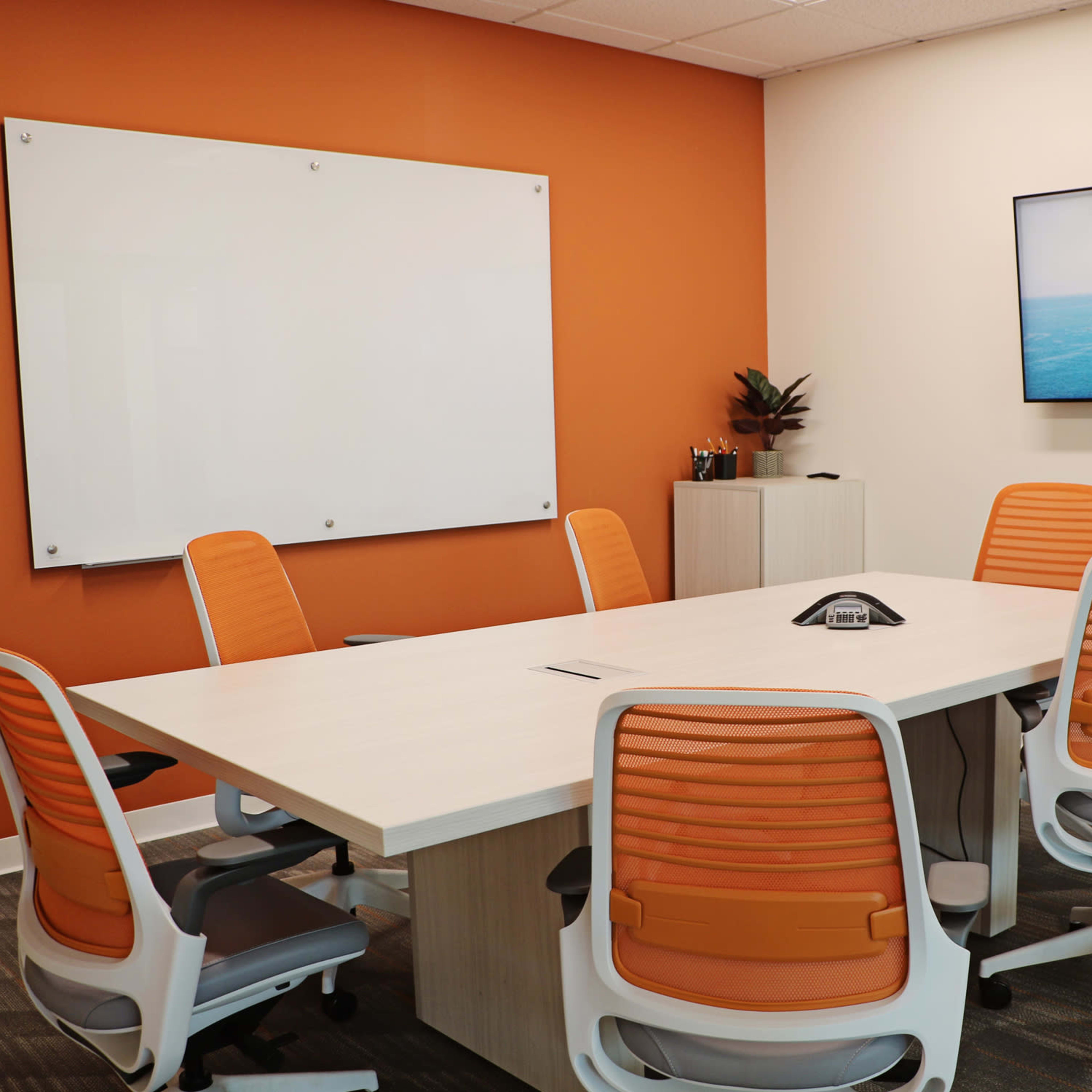 A modern conference room features a whiteboard, a wall-mounted TV displaying a calendar, a central table, and several orange and white chairs.