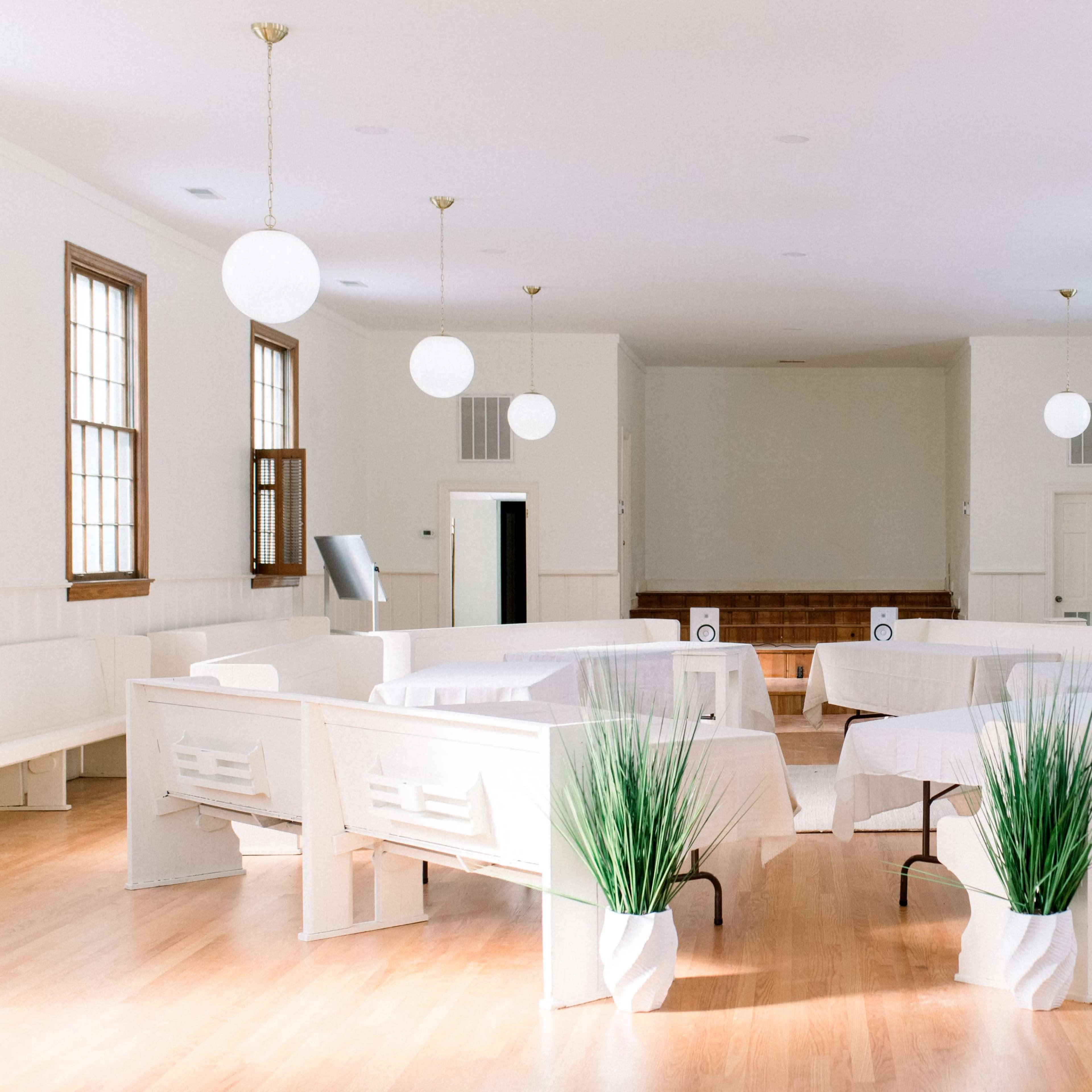 A spacious, well-lit room with white walls, featuring round tables arranged in a circular layout and potted plants in white coverings.
