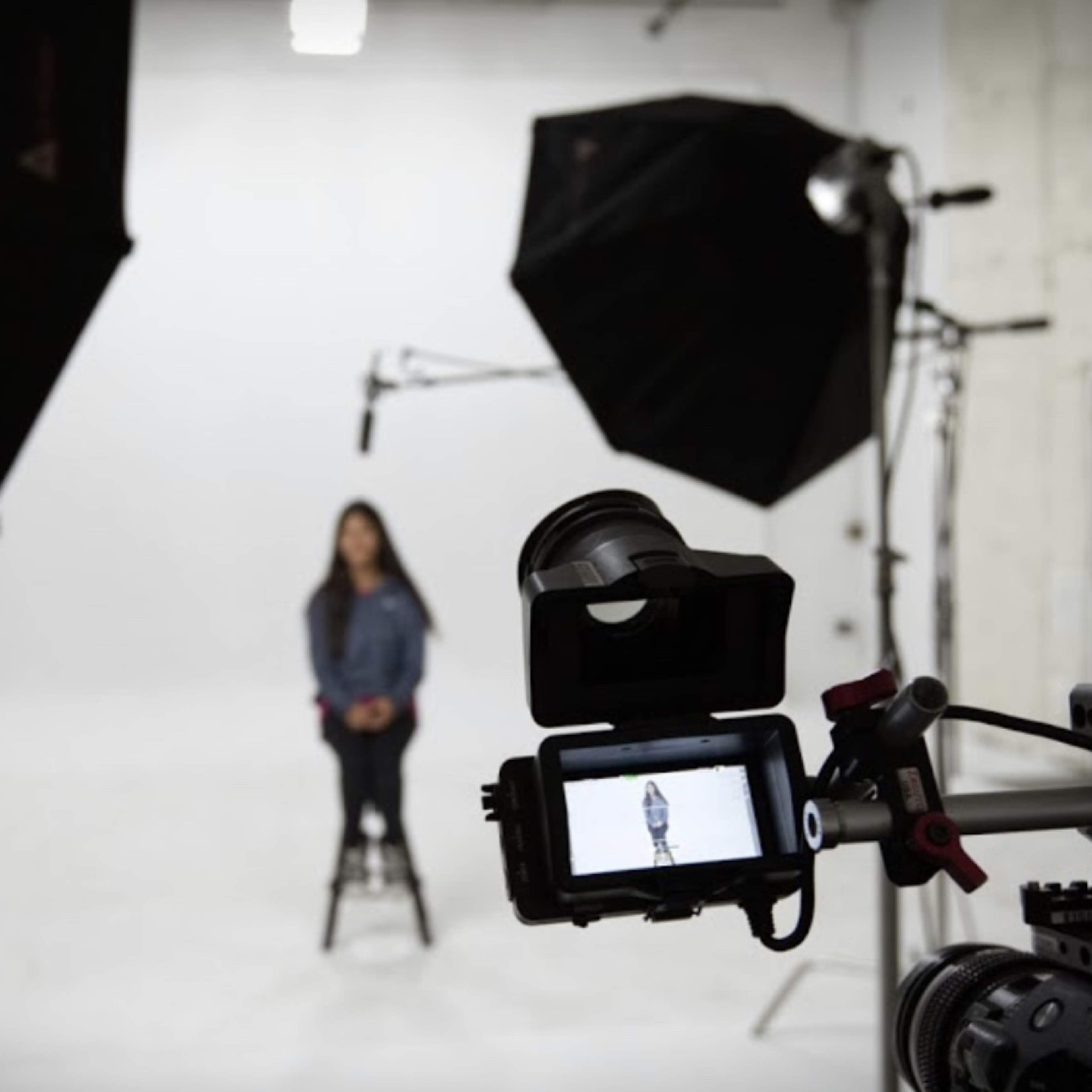 A video camera is positioned in front of a seated person in a brightly lit studio with softbox lights.