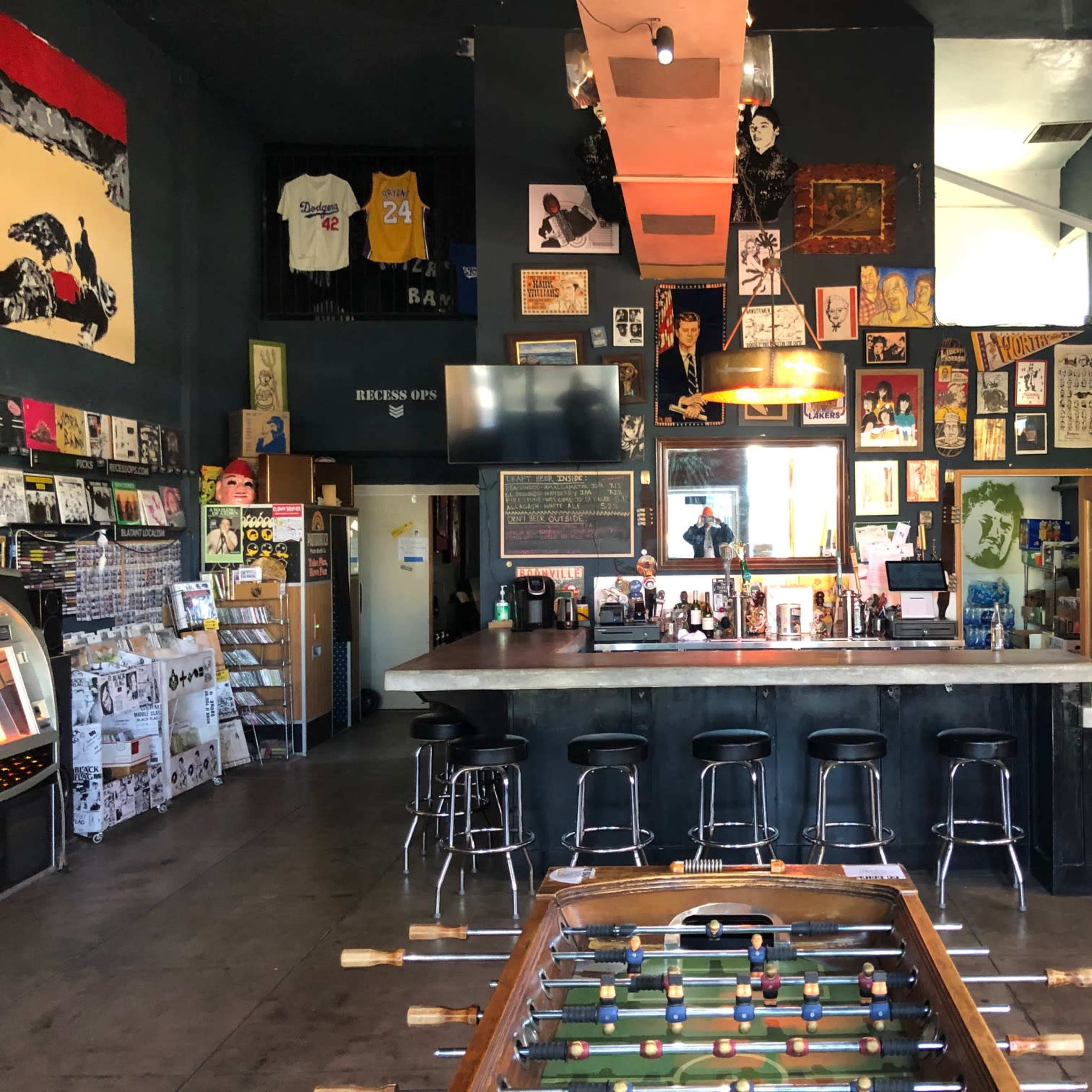 A dimly lit bar with a foosball table in the foreground, surrounded by various posters and artwork on the walls.