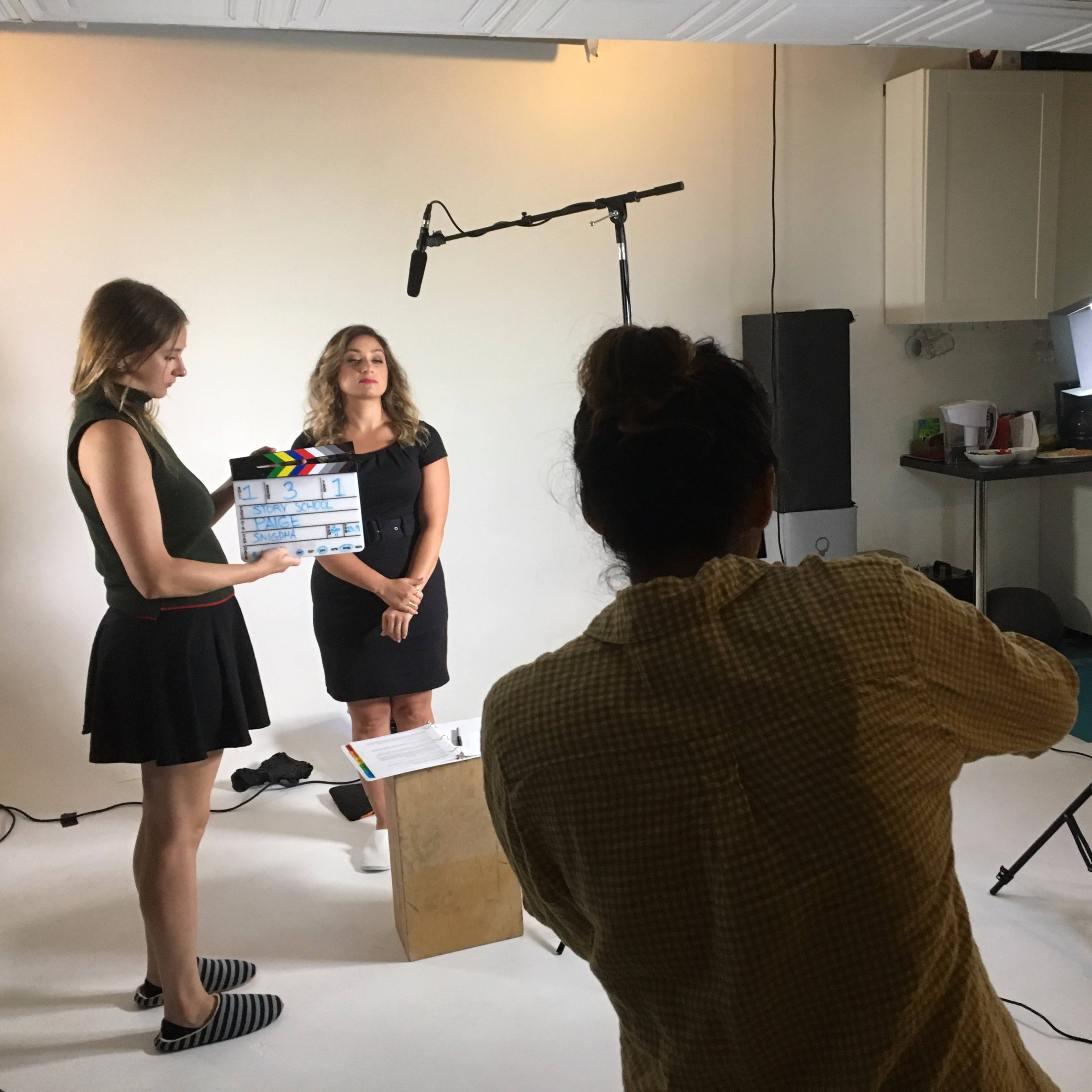 A woman holding a clapperboard prepares to film another woman in front of a camera in a well-lit studio setting.